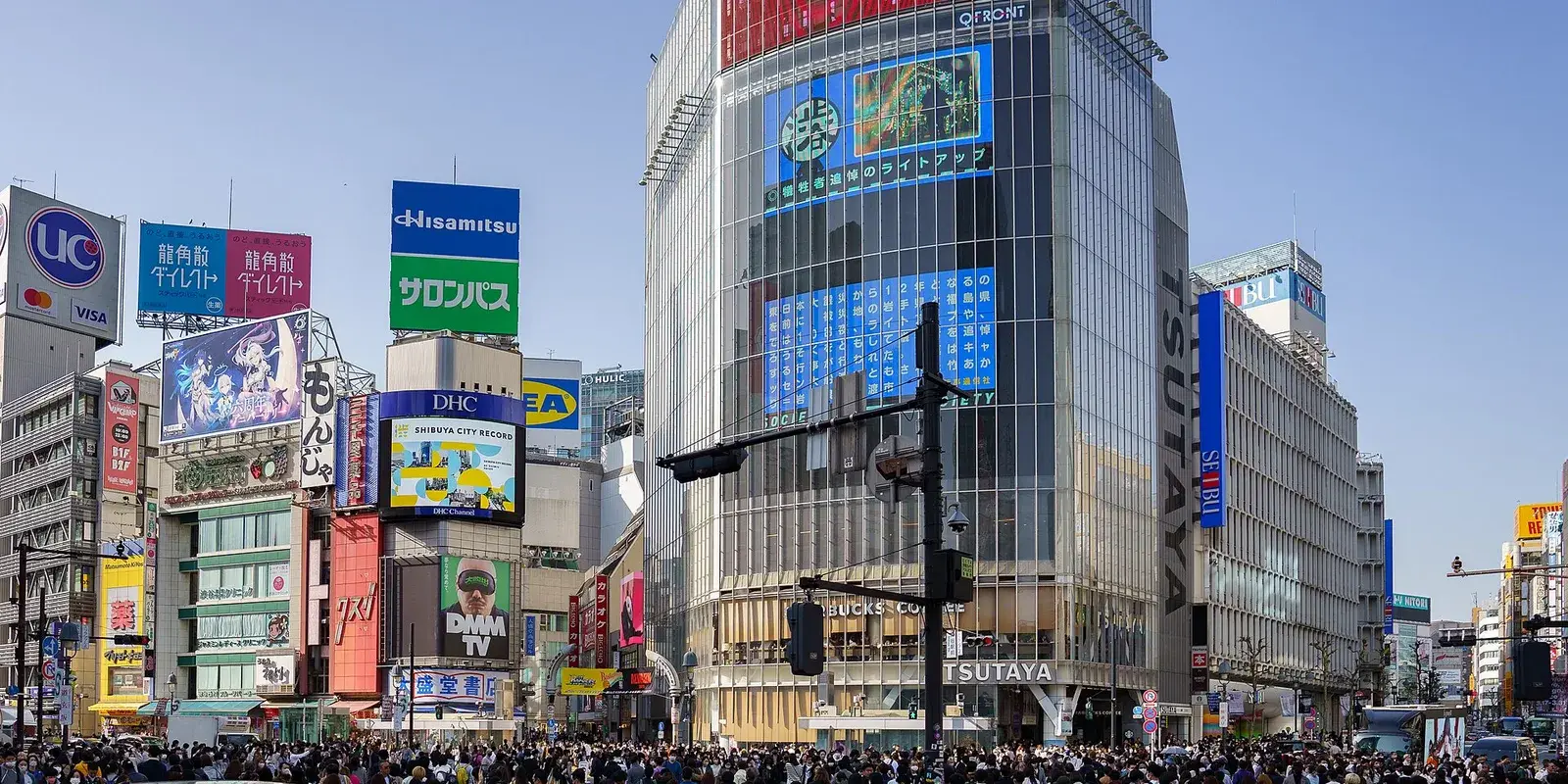 Crowds of pedestrians filling the Shibuya Scramble Crossing in Tokyo on a clear day, with surrounding billboards and buildings