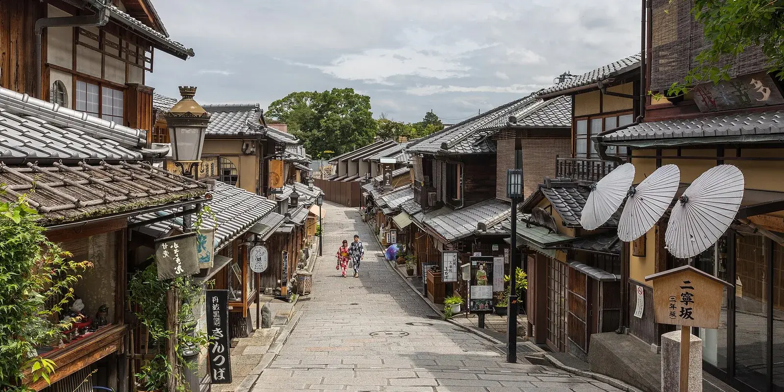 Travelers in yukata walk up the traditional stone-paved Ninenzaka street in Higashiyama, Kyoto, lined with wooden machiya houses and paper umbrellas