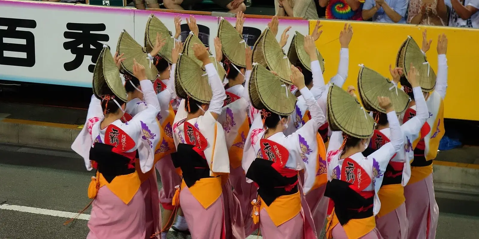 A row of Awa Odori dancers seen from behind in deep conical straw amigasa hats and pink kimono, arms raised in the festival's signature pose, along a lantern-lit street in Tokushima