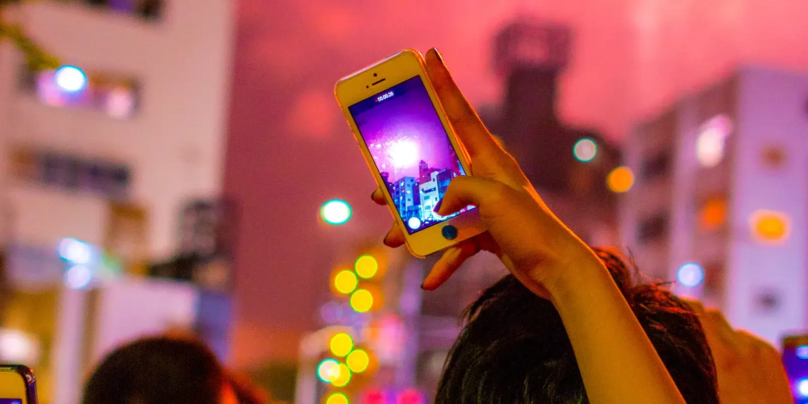A hand raises a smartphone to film fireworks over a Tokyo street at night, the live camera view glowing on its screen above a crowd seen only from behind