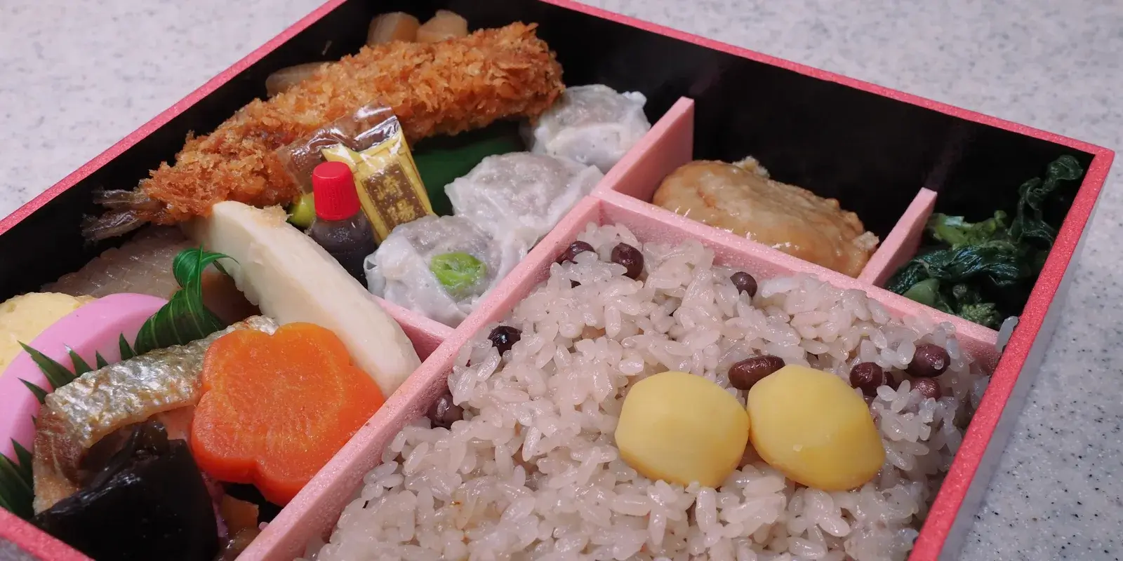 A Japanese boxed meal (bento) of red-bean sekihan rice, fried shrimp, shumai dumplings, simmered vegetables and chestnuts in a partitioned takeaway box