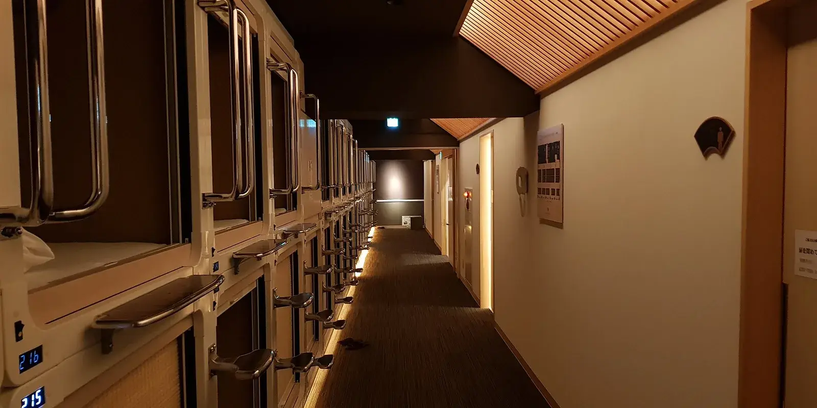 Warmly lit corridor of a Japanese capsule hotel lined with stacked sleeping pods, with no people present