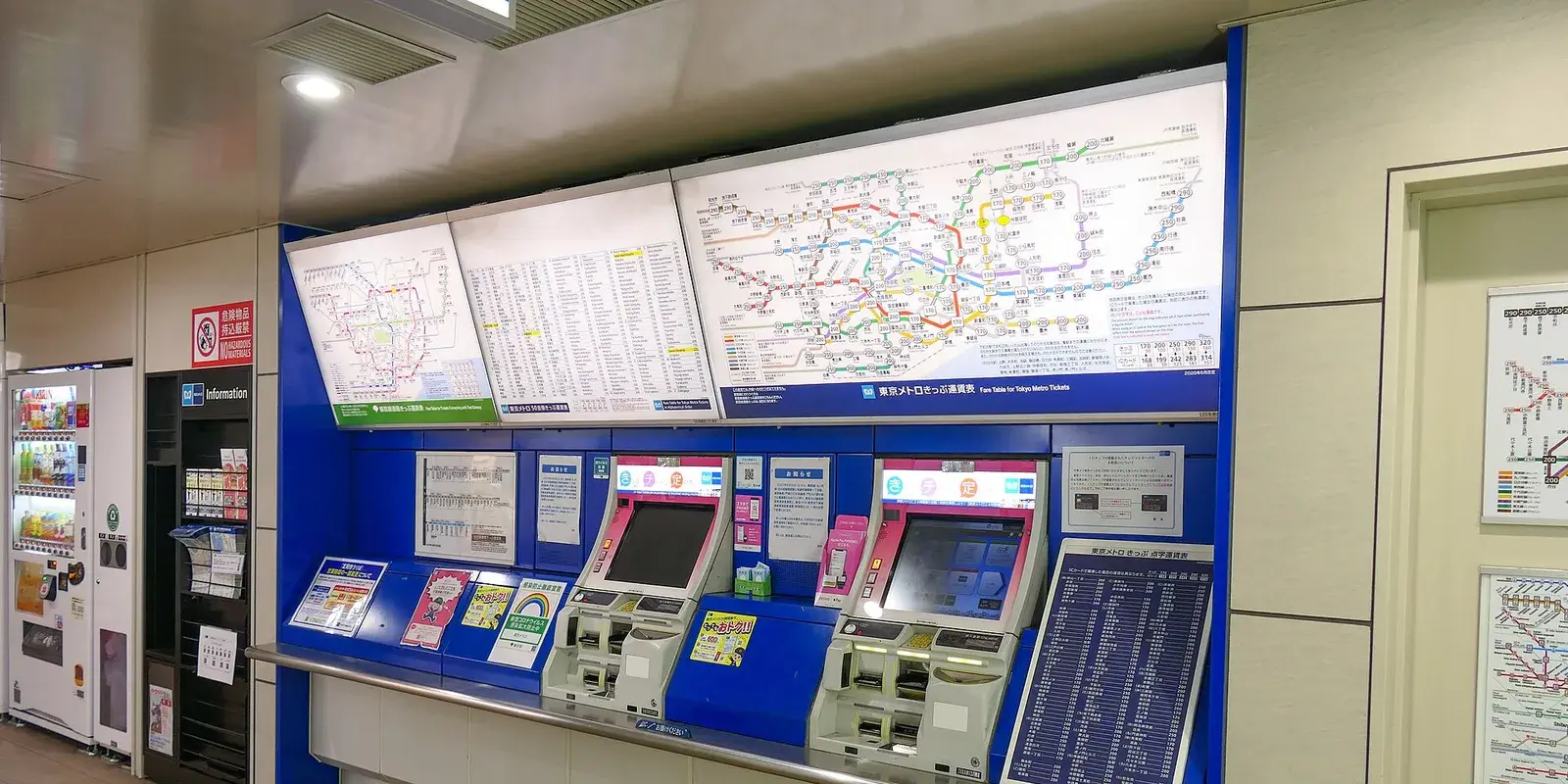 A wall of colorful Tokyo subway route maps and kanji fare tables above ticket vending machines at Ueno-Hirokoji Station, with no people present