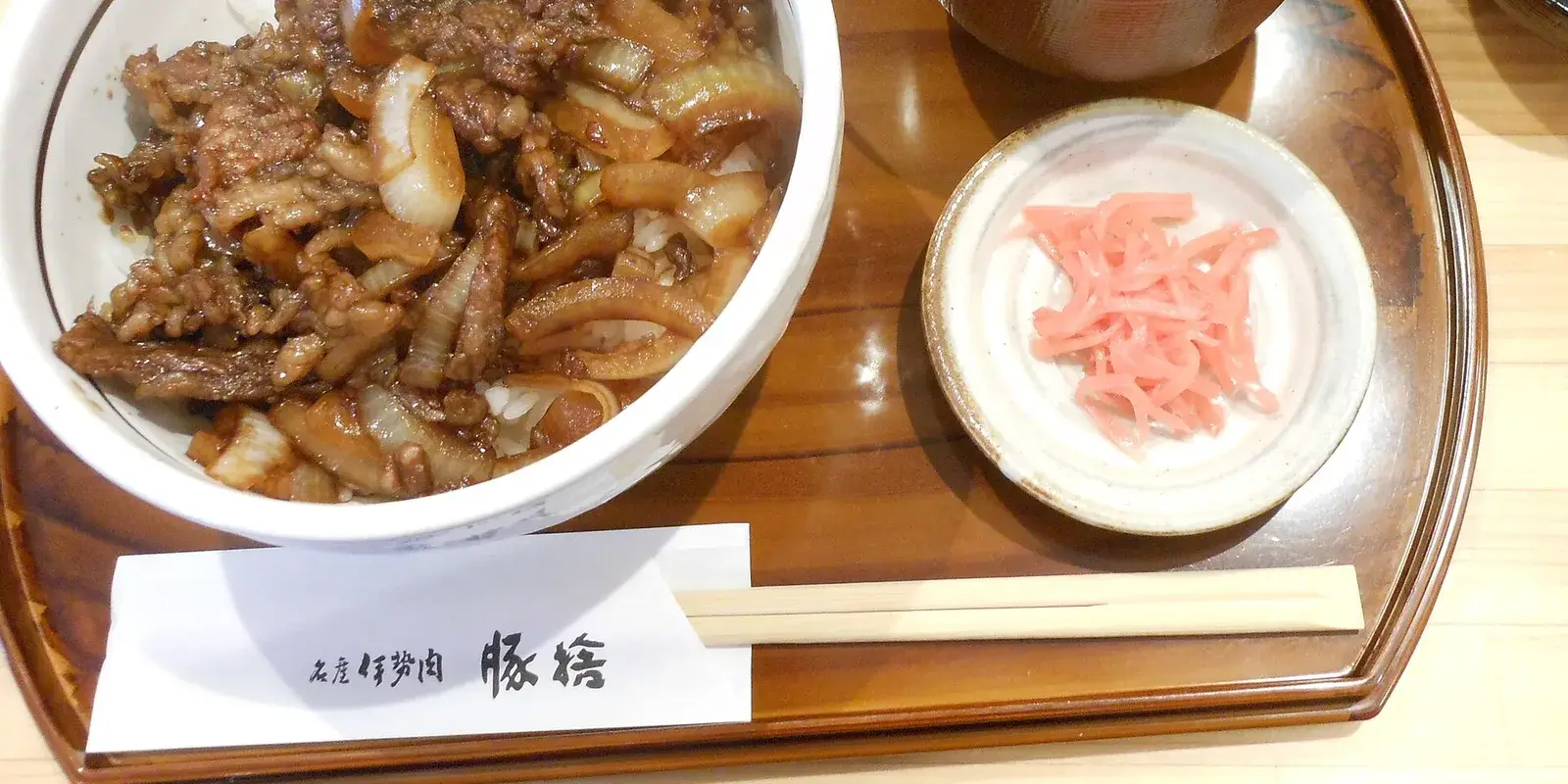 A Japanese gyudon (beef bowl) set meal on a wooden tray, with miso soup, pickled ginger, and wooden chopsticks resting on a paper sleeve