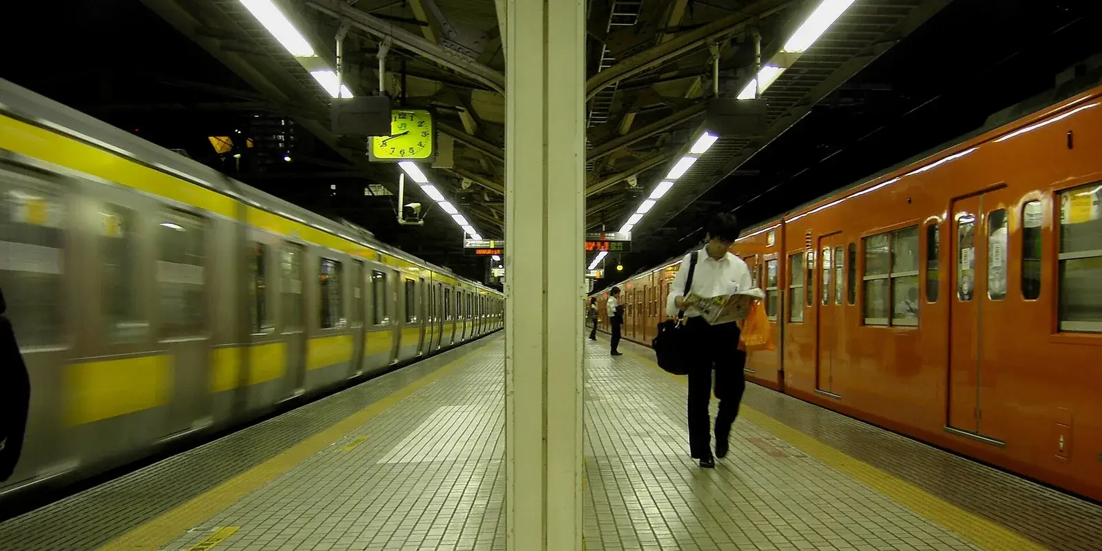 A Japanese commuter train platform in Tokyo at night, with a yellow Sobu Line train and an orange Chuo Line train on either side and a lone office worker walking with a bag