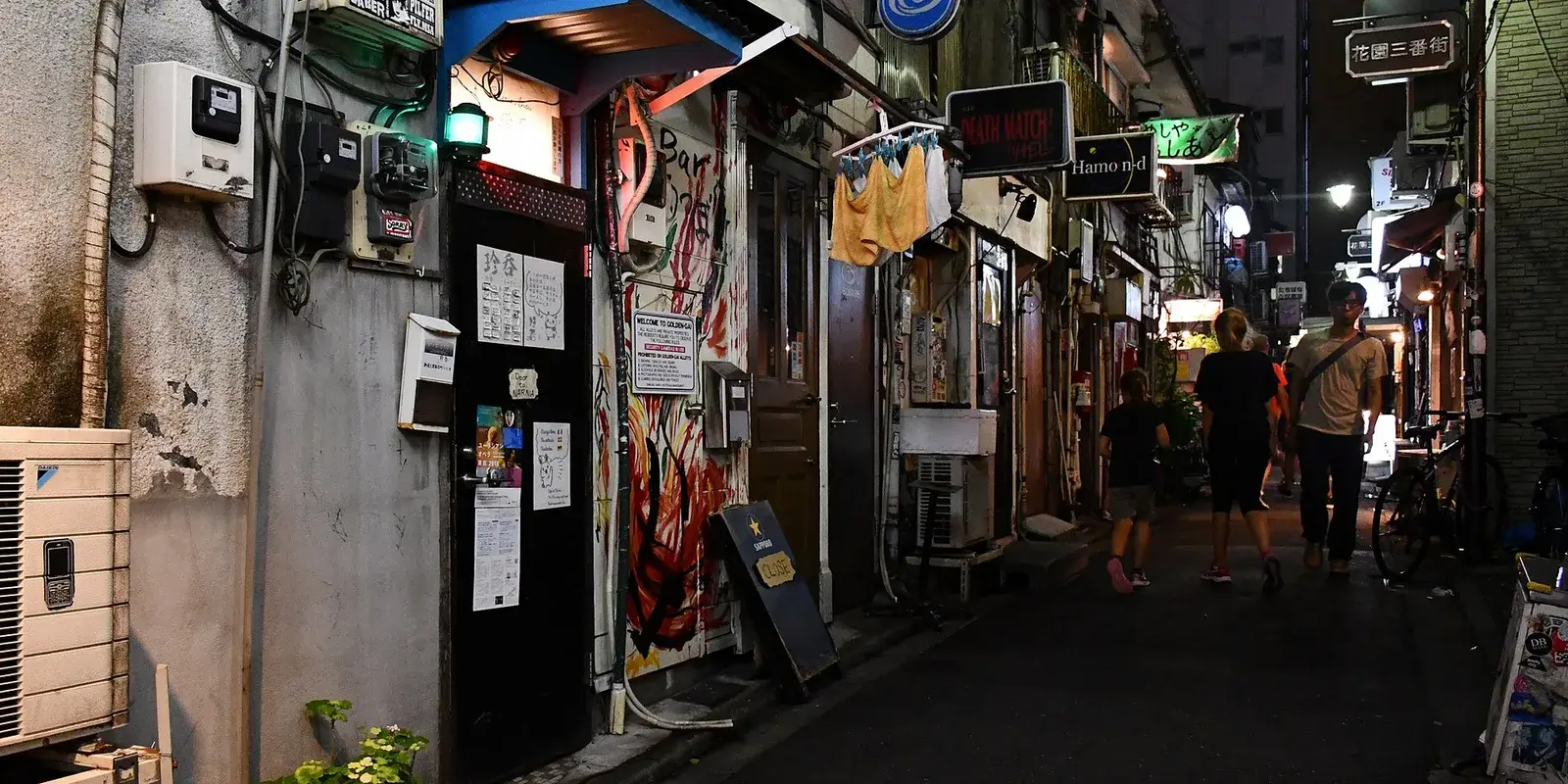 A narrow Golden Gai alley in Shinjuku at night, lined with tiny shanty-style bars, glowing signs and a lantern, with a few distant figures down the lane