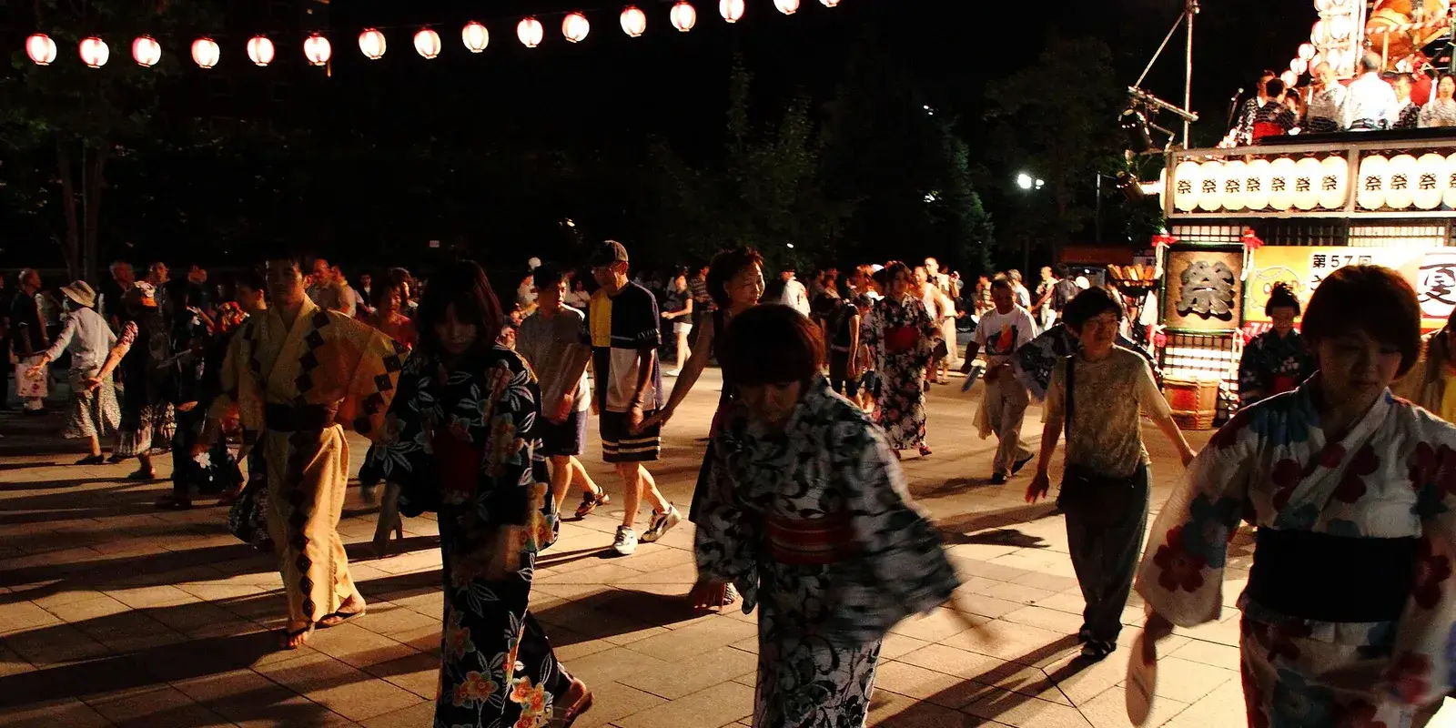 Yukata-clad dancers in a bon odori circle at a nighttime Japanese summer festival, with paper lanterns strung overhead and a lit yagura tower