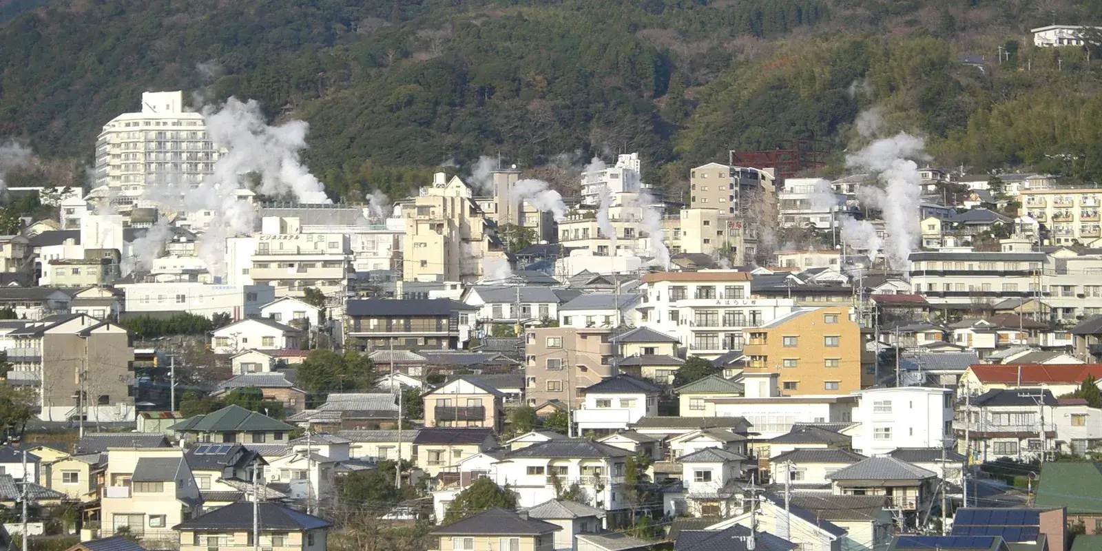 Columns of white steam rising from the rooftops of Beppu's Kannawa district, with the bare winter slopes of Mount Tsurumi behind