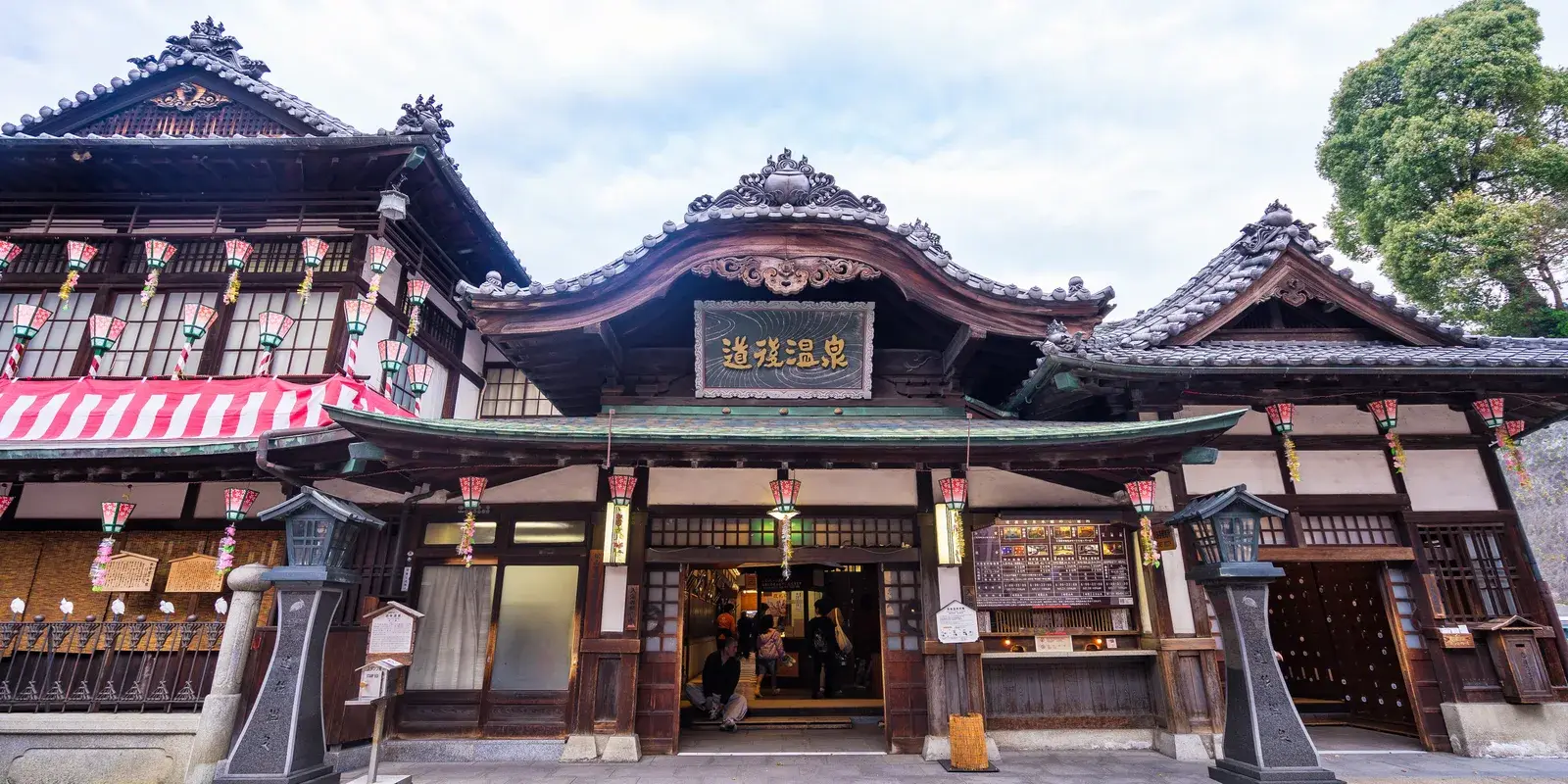 The Dogo Onsen Honkan, a three-storey wooden public bathhouse with carved gables and the Dogo Onsen sign, in daylight, Matsuyama