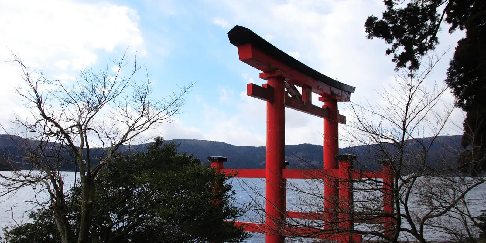 The red torii gate of Hakone Shrine rising from Lake Ashinoko under a partly clouded sky, framed by bare branches
