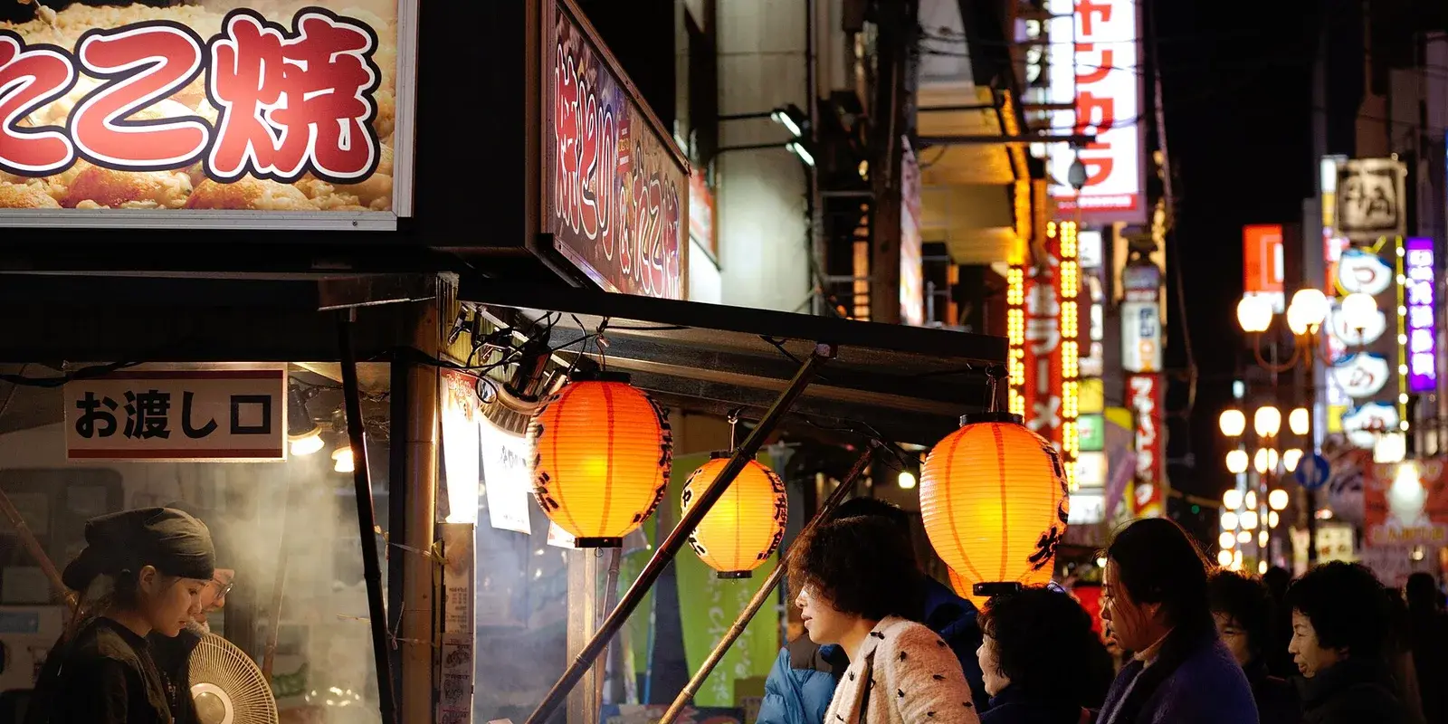 Takoyaki street-food stall with glowing red lanterns and neon signs on a busy Dotonbori food street at night, with people gathering to buy snacks