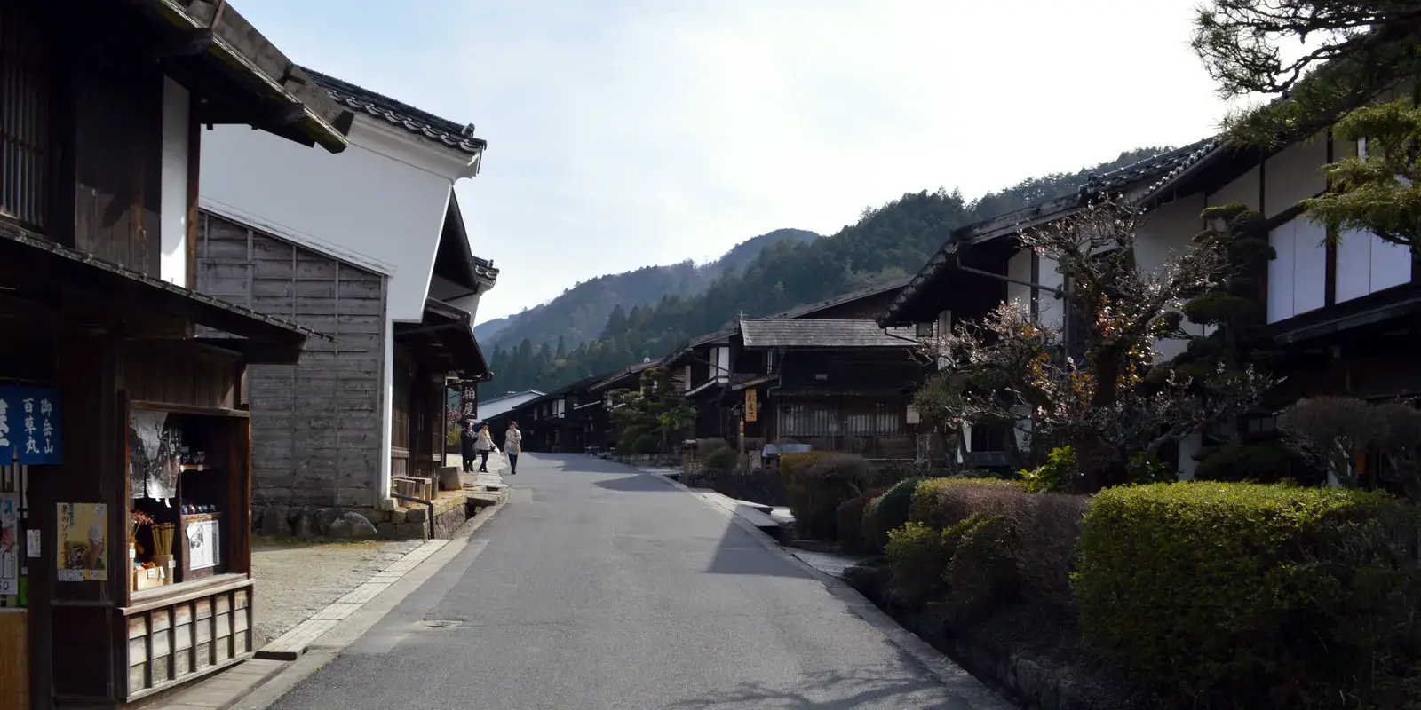 A quiet street of wooden post-town houses in Tsumago-juku, Nagano, curving toward forested mountains, with a few distant walkers and a blossoming tree
