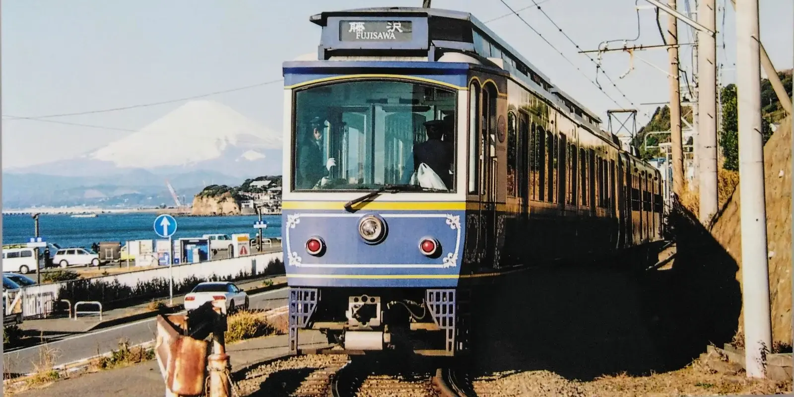 A little green-and-cream Enoden train running along the Shonan coast, with Mount Fuji across the bay near Kamakura