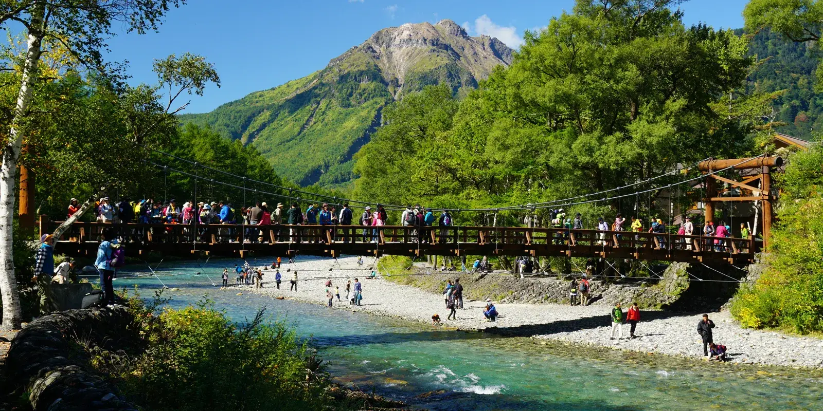 Visitors crossing the wooden Kappa Bridge over the turquoise Azusa River in Kamikochi, with Mount Yake rising behind under a clear sky