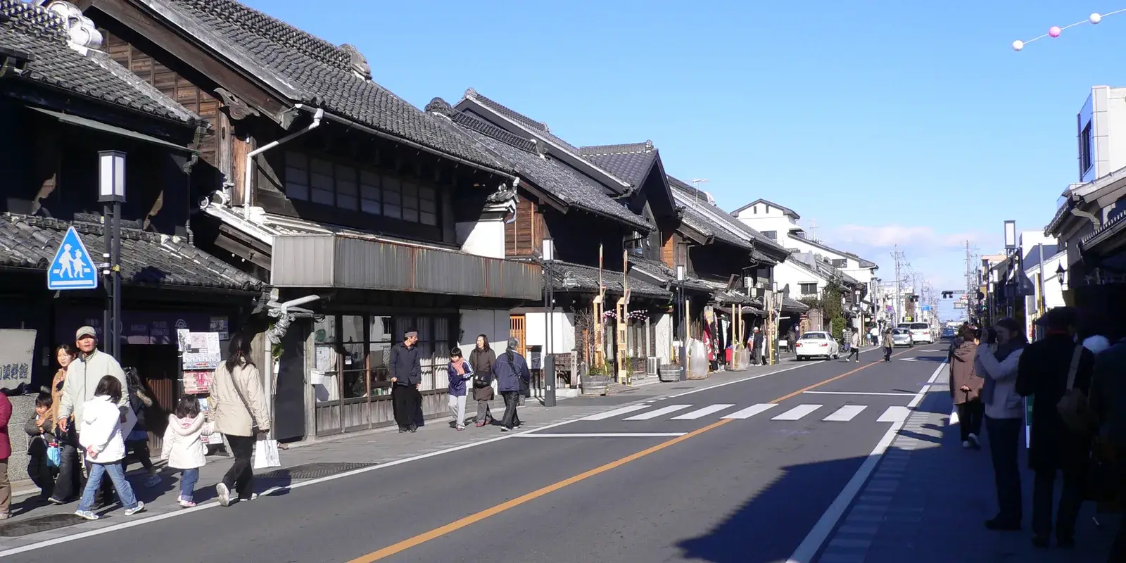 A clear-sky view down Kurazukuri Street in Kawagoe, dark wooden warehouse buildings lining a road with pedestrians and a pedestrian crossing