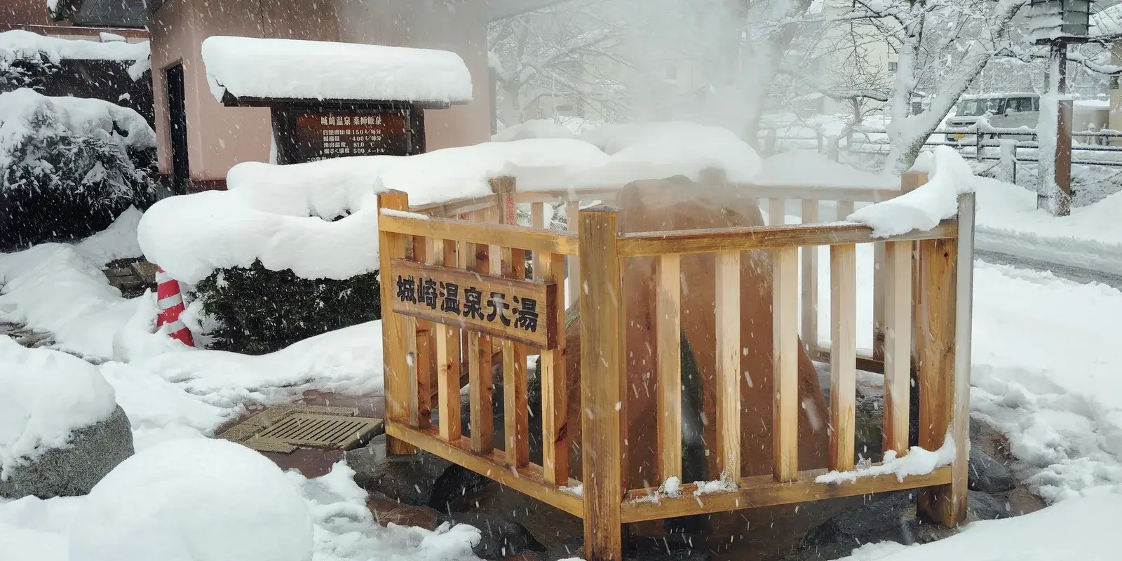 The Motoyu hot-spring source in Kinosaki Onsen, steam rising from the spring behind a wooden railing as snow falls
