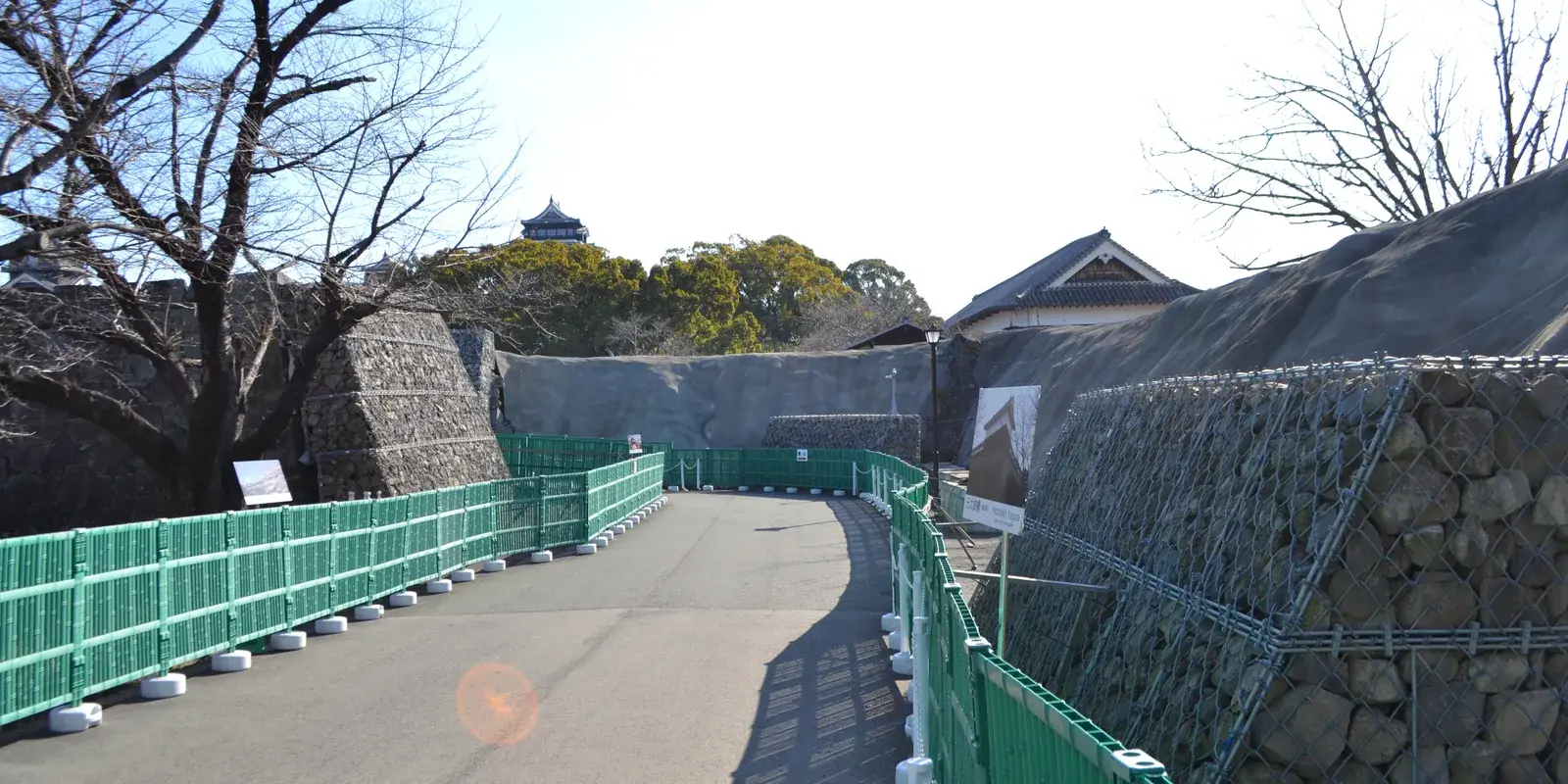 The approach path through Kumamoto Castle's grounds, its earthquake-damaged stone walls held in wire cages for repair, with the reconstructed main keep beyond