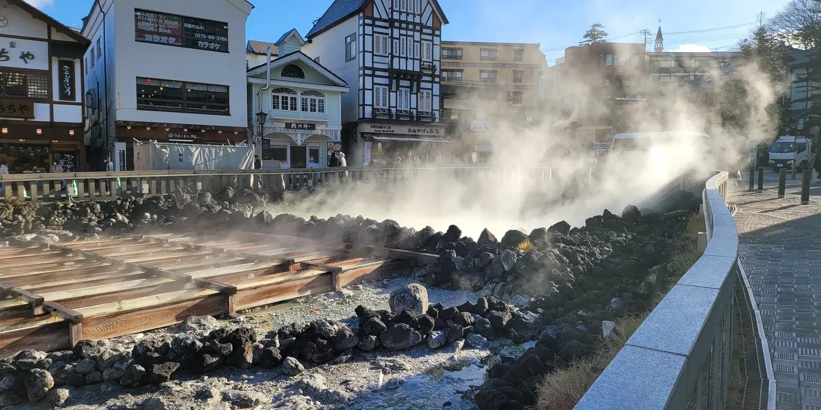 Steam rising off the wooden cooling channels of Yubatake in Kusatsu Onsen on a clear day, ryokan and shop buildings lining the street beyond