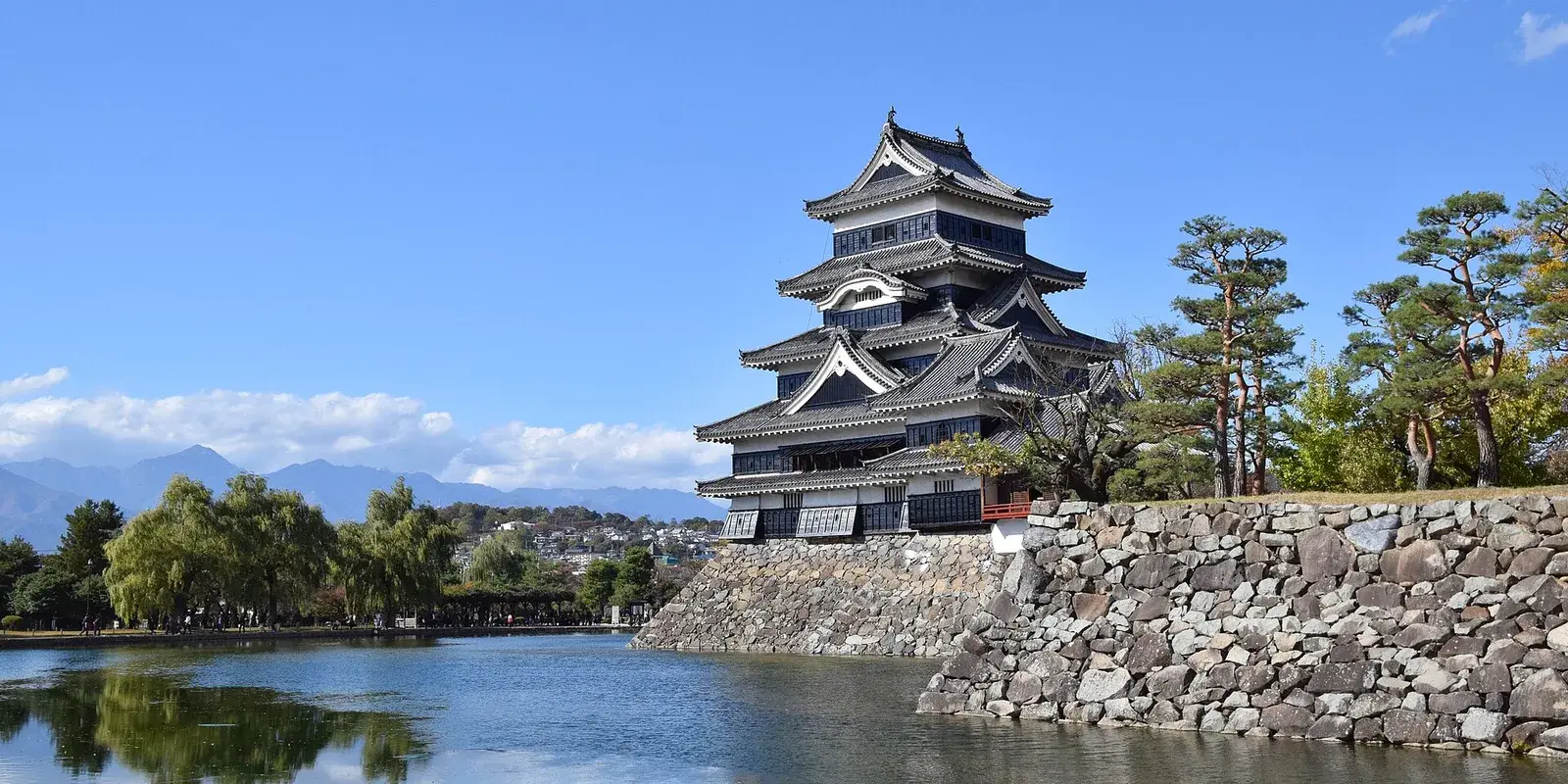 The black keep of Matsumoto Castle reflected in its moat, with the Northern Alps on the horizon under a clear autumn sky