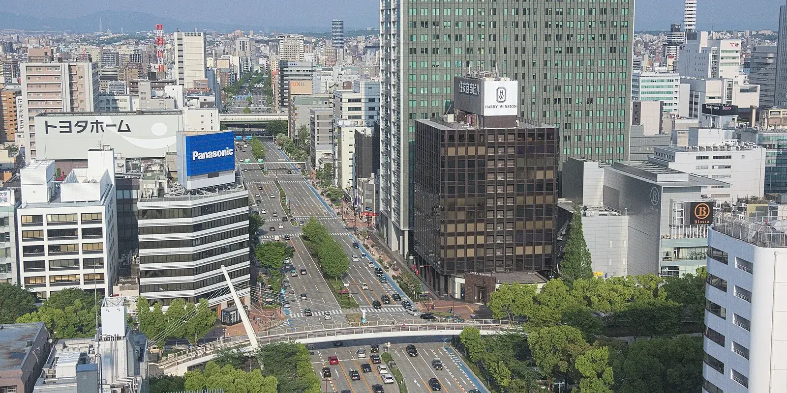 An elevated view down the wide Sakae boulevard in central Nagoya, lined with office towers, corporate signage, and a footbridge crossing the intersection