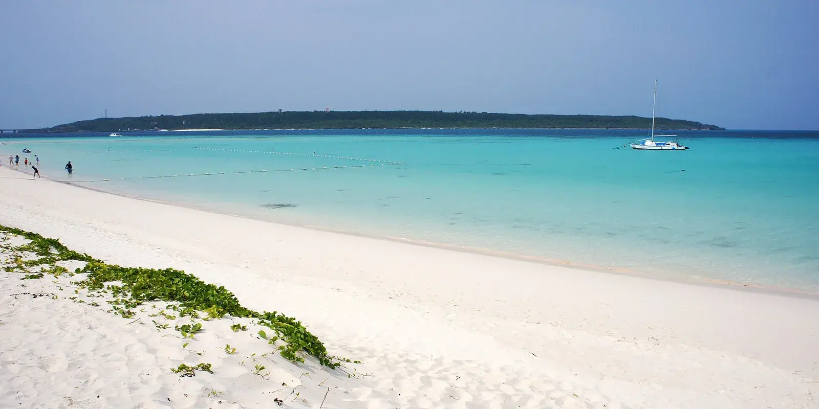 Yonaha Maehama, a long white-sand beach on Miyako Island, its shallows glowing the turquoise 'Miyako blue', with a small island and a sailboat on the horizon