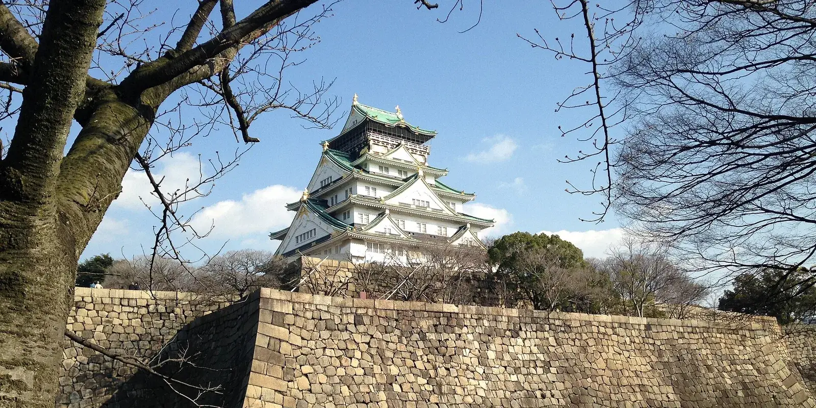 Osaka Castle main keep with its green roofs and gold trim rising above the massive Tokugawa-era stone wall, with the inner moat below