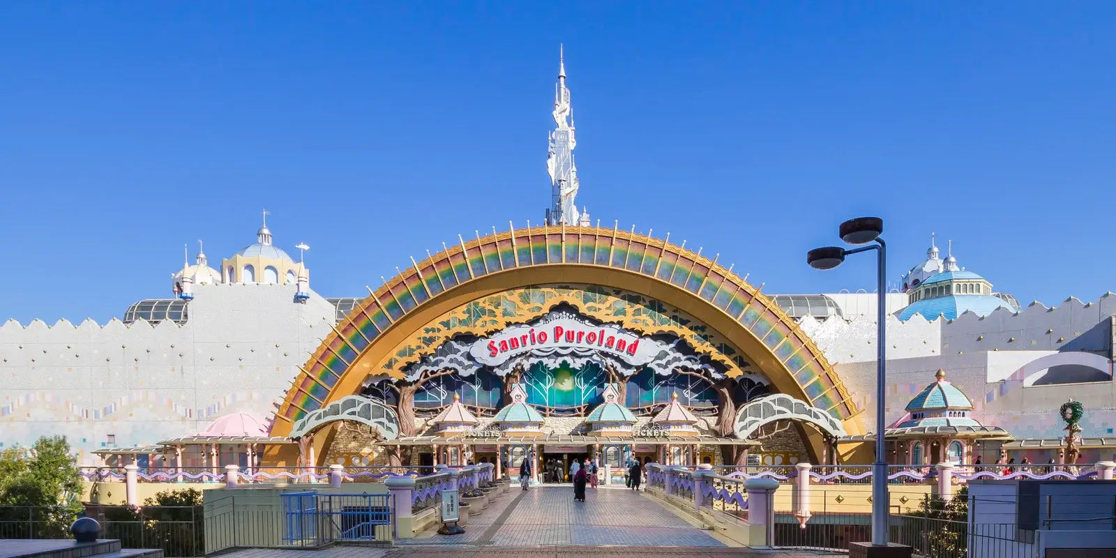 The rainbow-arched entrance of Sanrio Puroland against a clear blue sky, with the ticket booths below