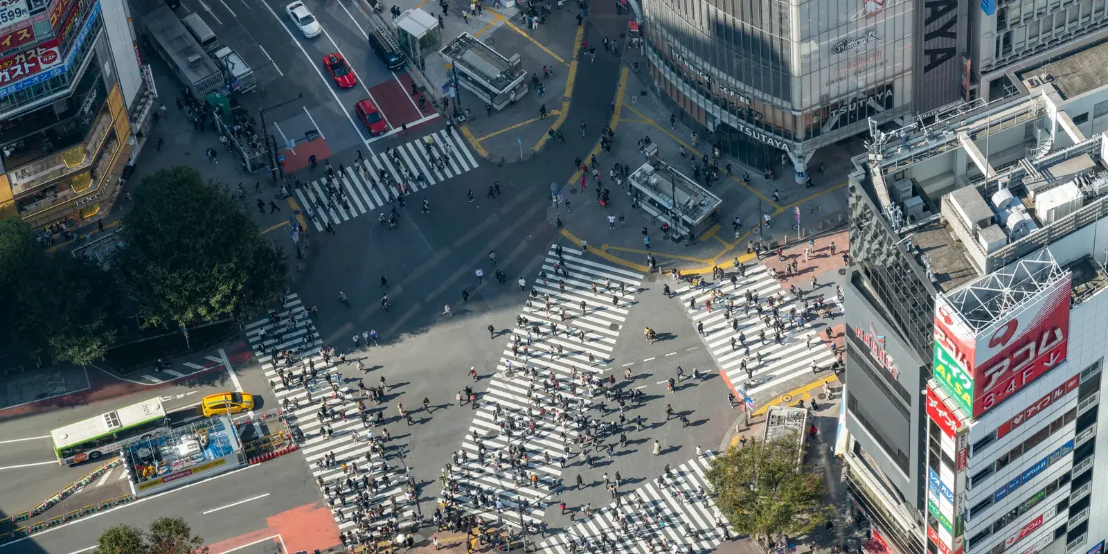 The Shibuya Scramble Crossing seen from above, its diagonal crosswalks filled with tiny figures, ringed by the buildings of central Shibuya