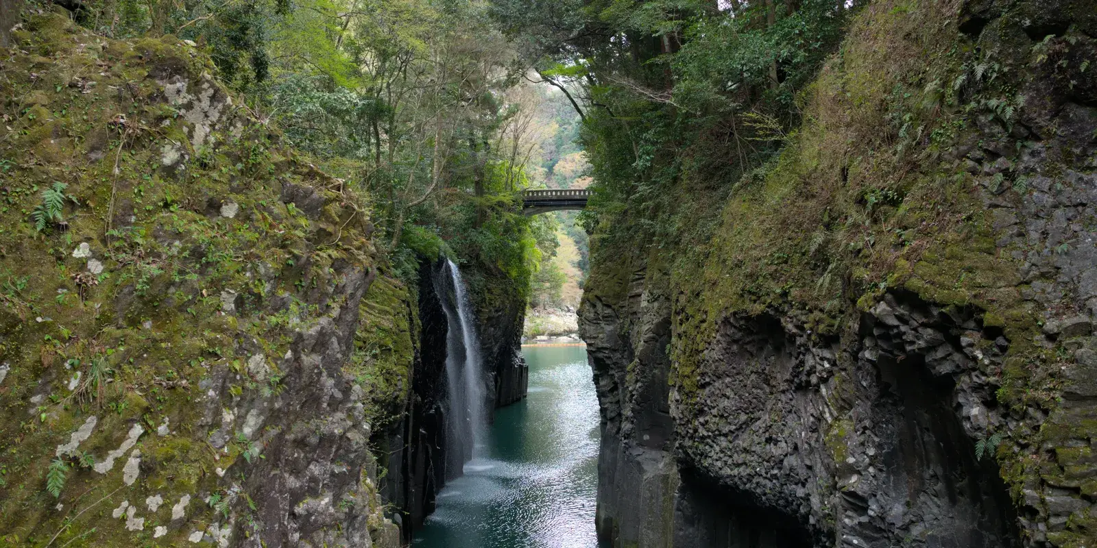 Manai Falls dropping into the emerald water of Takachiho Gorge, framed by columnar basalt cliffs, with an arch bridge beyond