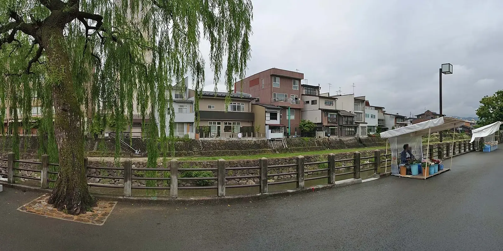 The willow-lined Miyagawa river in Takayama at a quiet hour, with a riverside morning-market stall setting up along the far bank