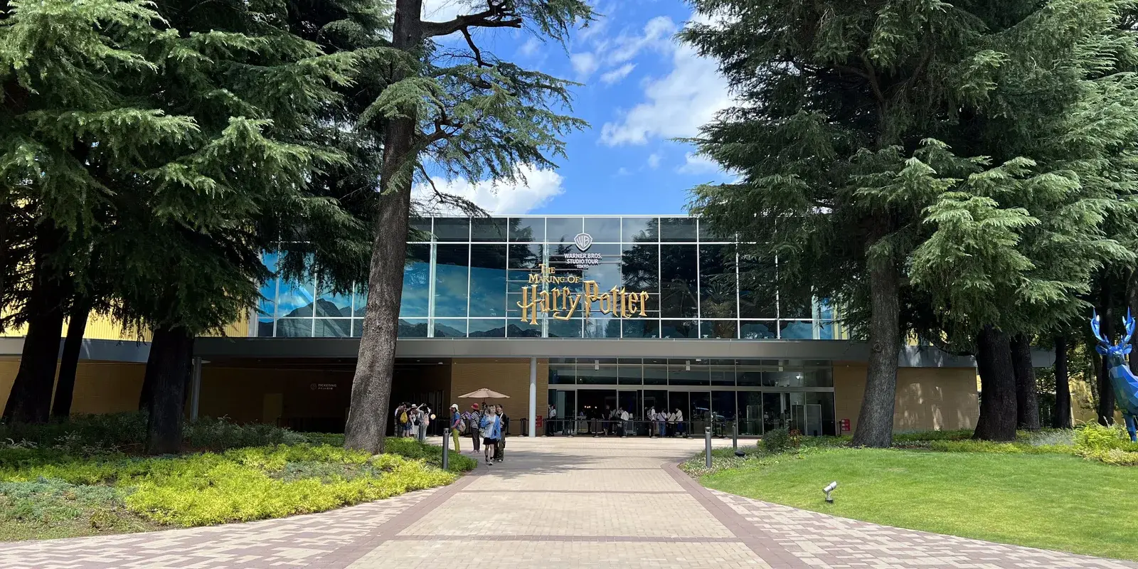 Visitors walk up the tree-lined path to the glass-fronted entrance of Warner Bros. Studio Tour Tokyo, with The Making of Harry Potter signage above the doors.