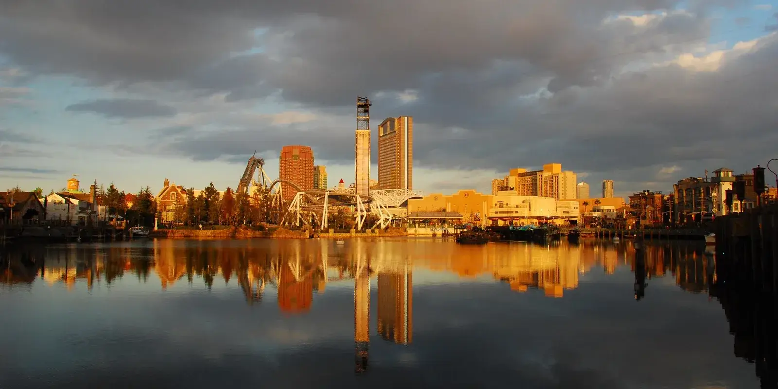 The Universal Studios Japan waterfront skyline reflected in the canal at golden hour, coasters and hotel towers lit warm against a cloudy sky
