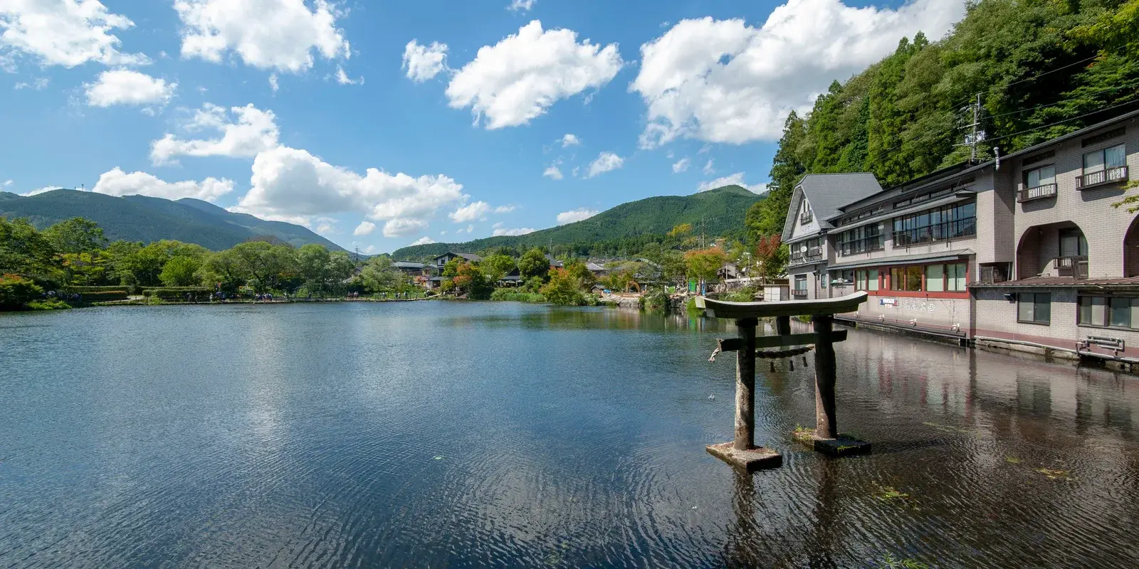 Lake Kinrin in Yufuin on a clear day, with a small stone torii standing in the shallow water and wooded hills behind