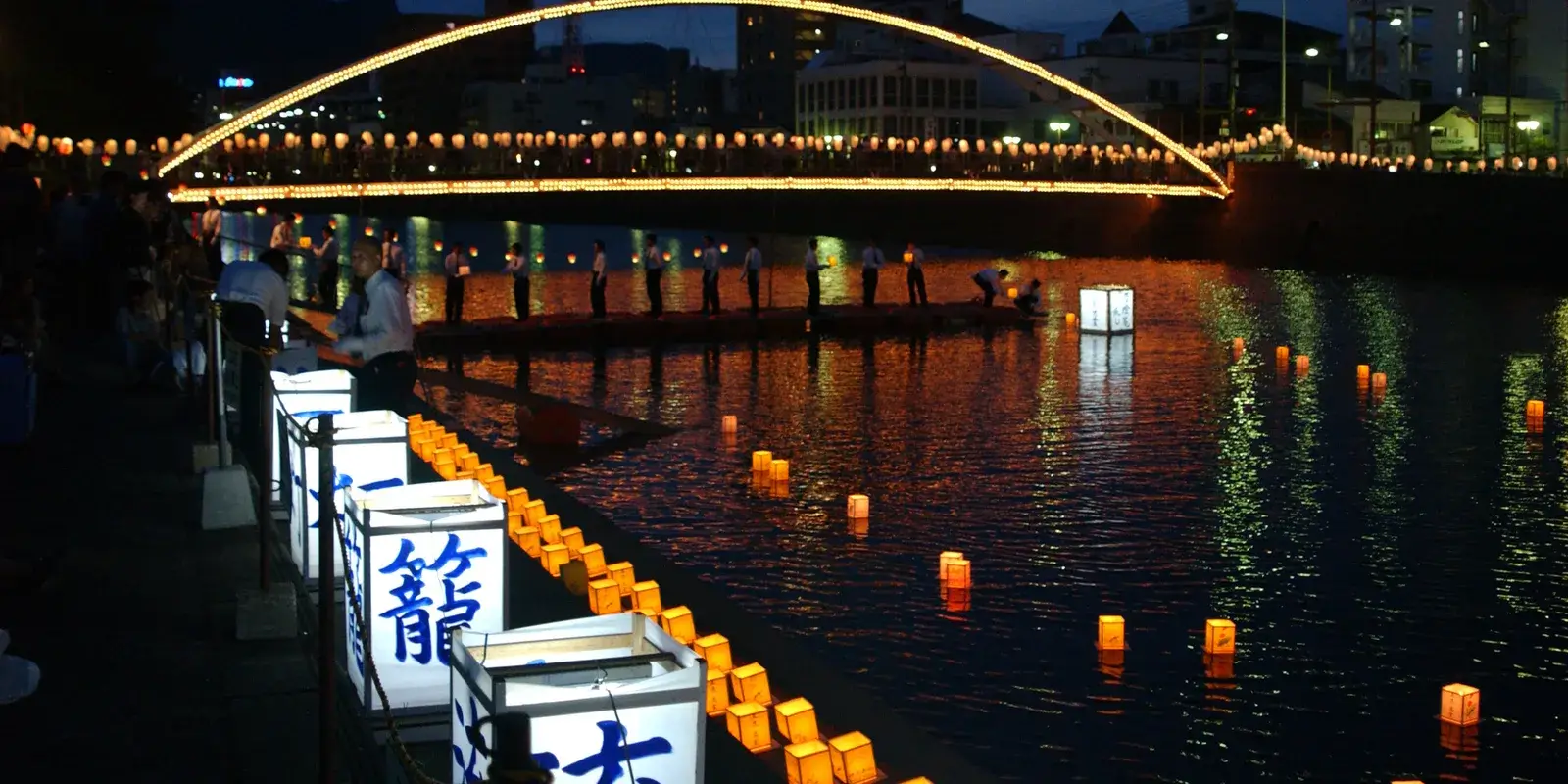 Glowing paper lanterns float on the Sasebo River beneath an illuminated bridge during the Obon festival at night