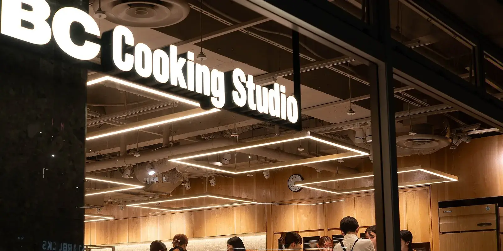 Storefront of an ABC Cooking Studio cooking school in Japan, its glowing sign above a warm wood-paneled kitchen where people gather for a class