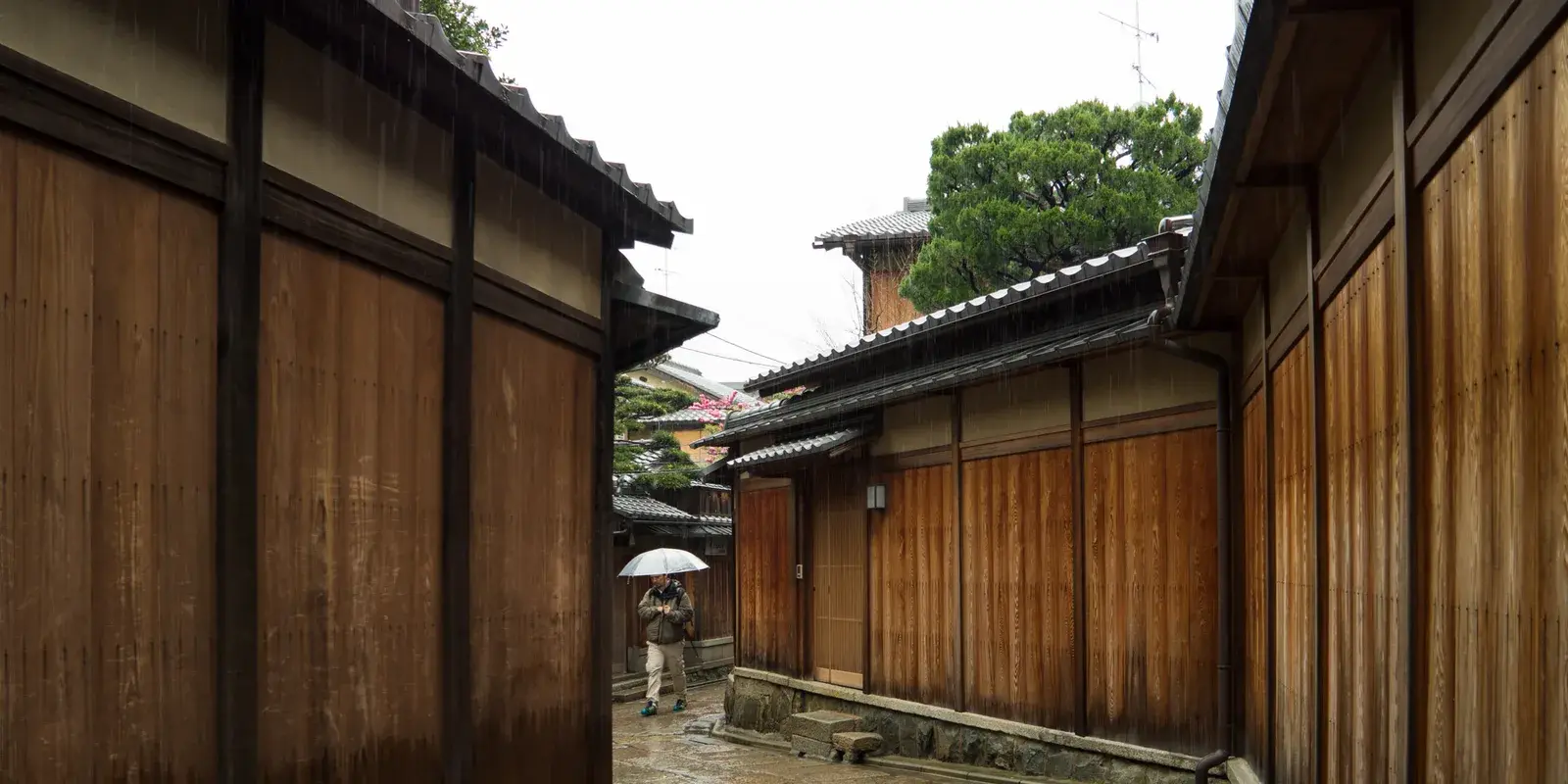 A quiet rainy alley in Kyoto lined with traditional wooden machiya walls and wet stone pavement, with a distant figure under an umbrella