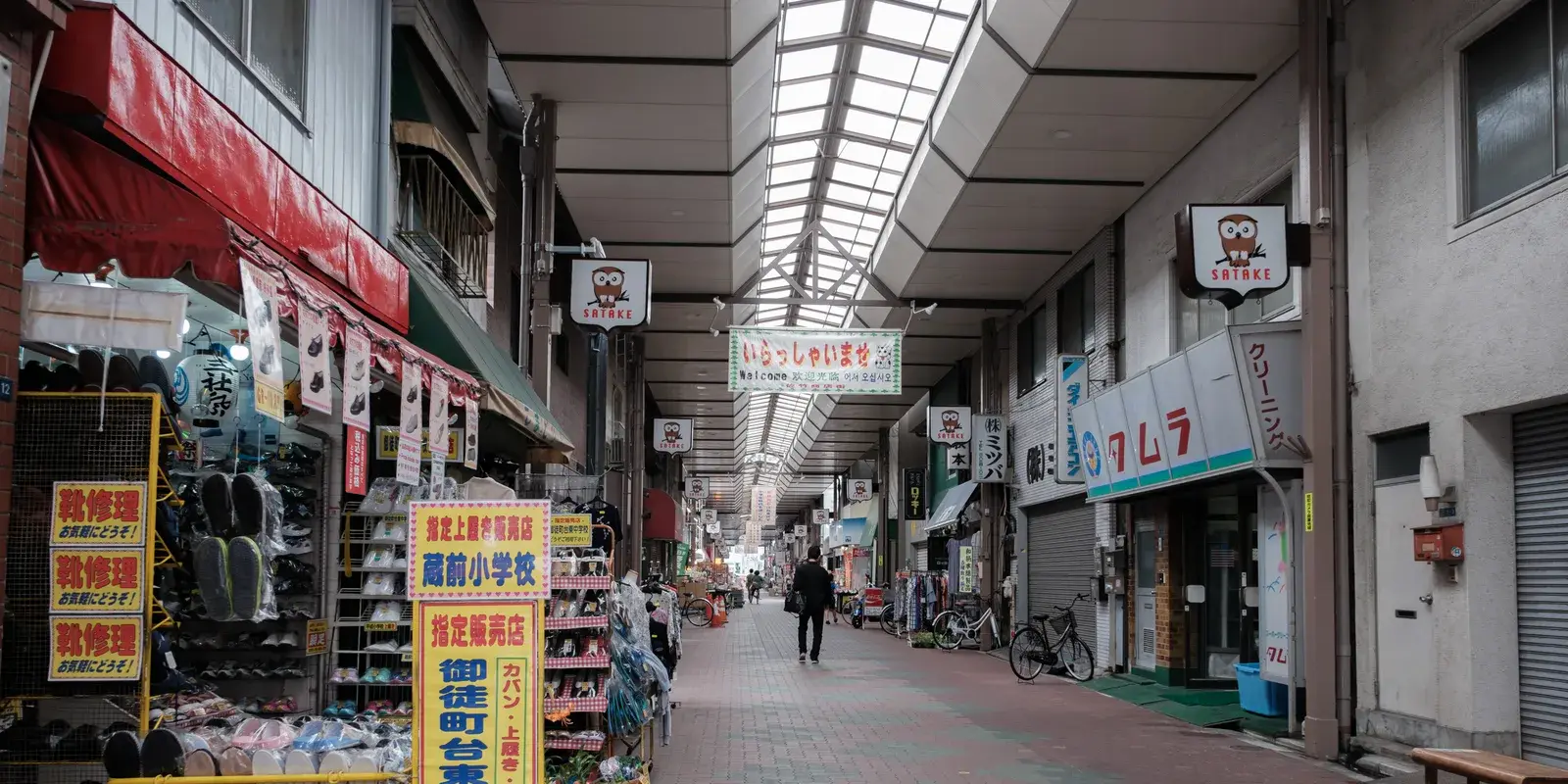 A covered local shopping street (shotengai) in a Tokyo neighborhood, with shops lining both sides and a single distant person walking away under the arcade roof