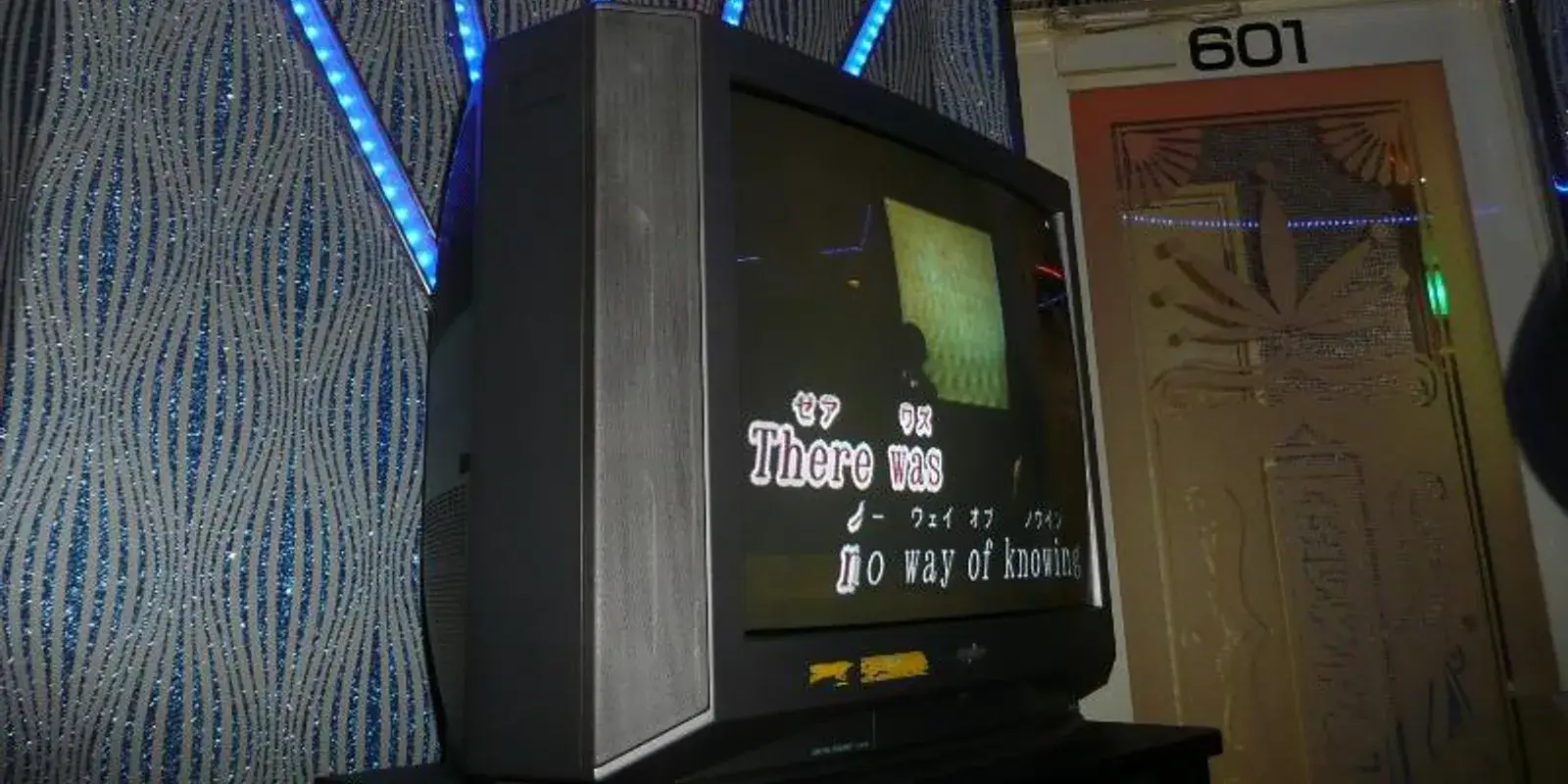 Interior of a Japanese karaoke box, with English and Japanese song lyrics on the screen, blue LED accent lighting, and the room door numbered 601