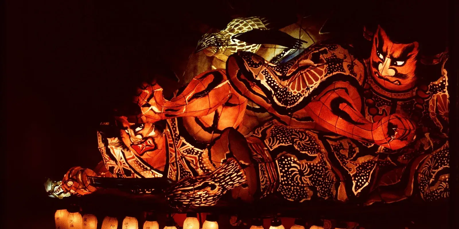 An illuminated Aomori Nebuta warrior float glowing against the night sky above a row of lit paper lanterns