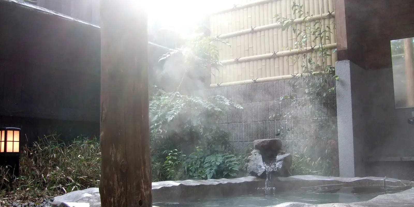 A steaming outdoor stone onsen bath (rotenburo) with a rock water spout, bamboo fence, and stone lantern at a Japanese hot spring