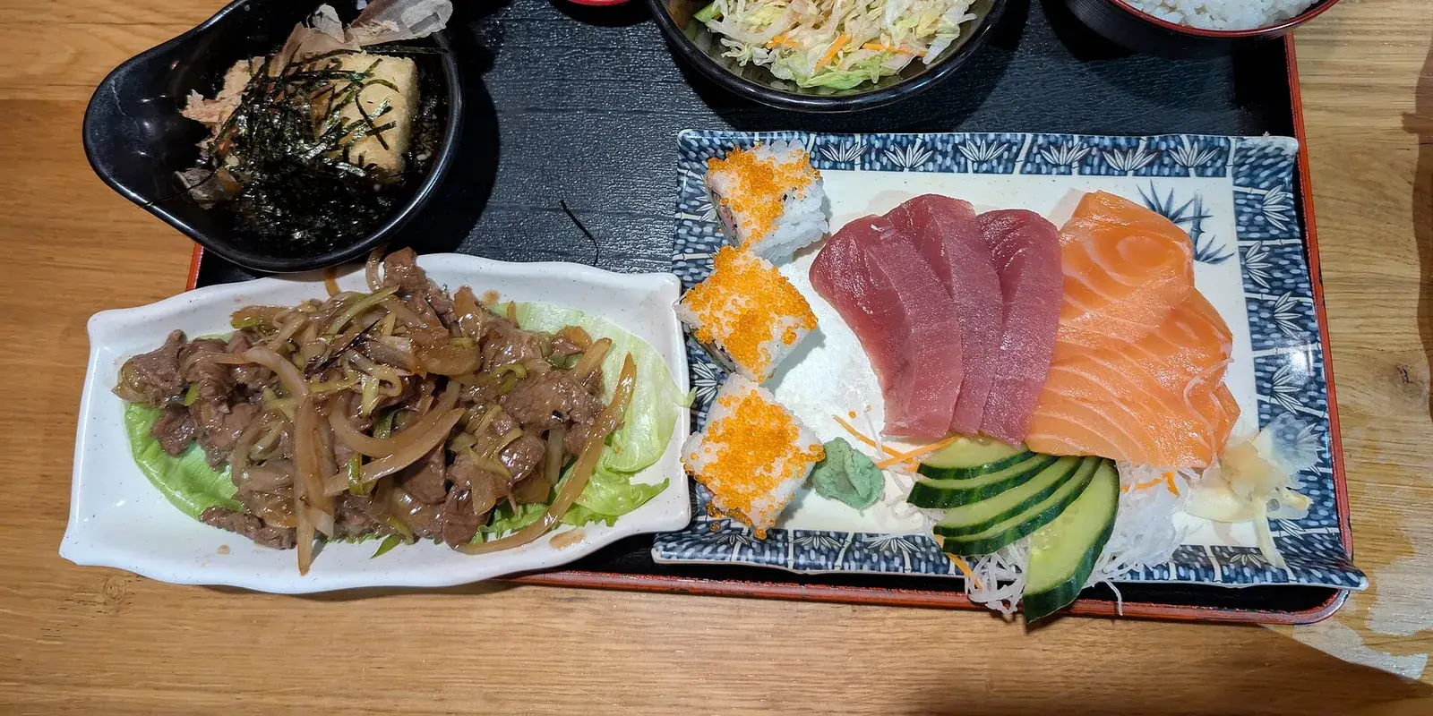 A Japanese teishoku set meal on a wooden table: sliced sashimi on a blue-and-white plate, a simmered side dish, salad, a bowl of rice, and miso soup