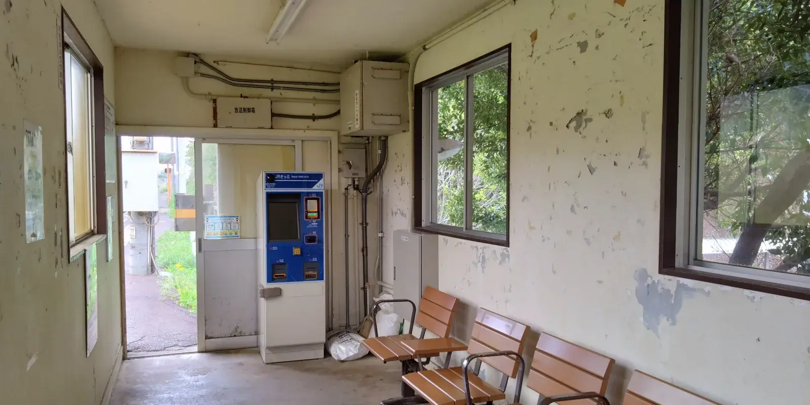 An empty, weathered rural Japanese train station waiting room with peeling walls, vacant wooden benches, a single ticket machine, and greenery visible through the windows