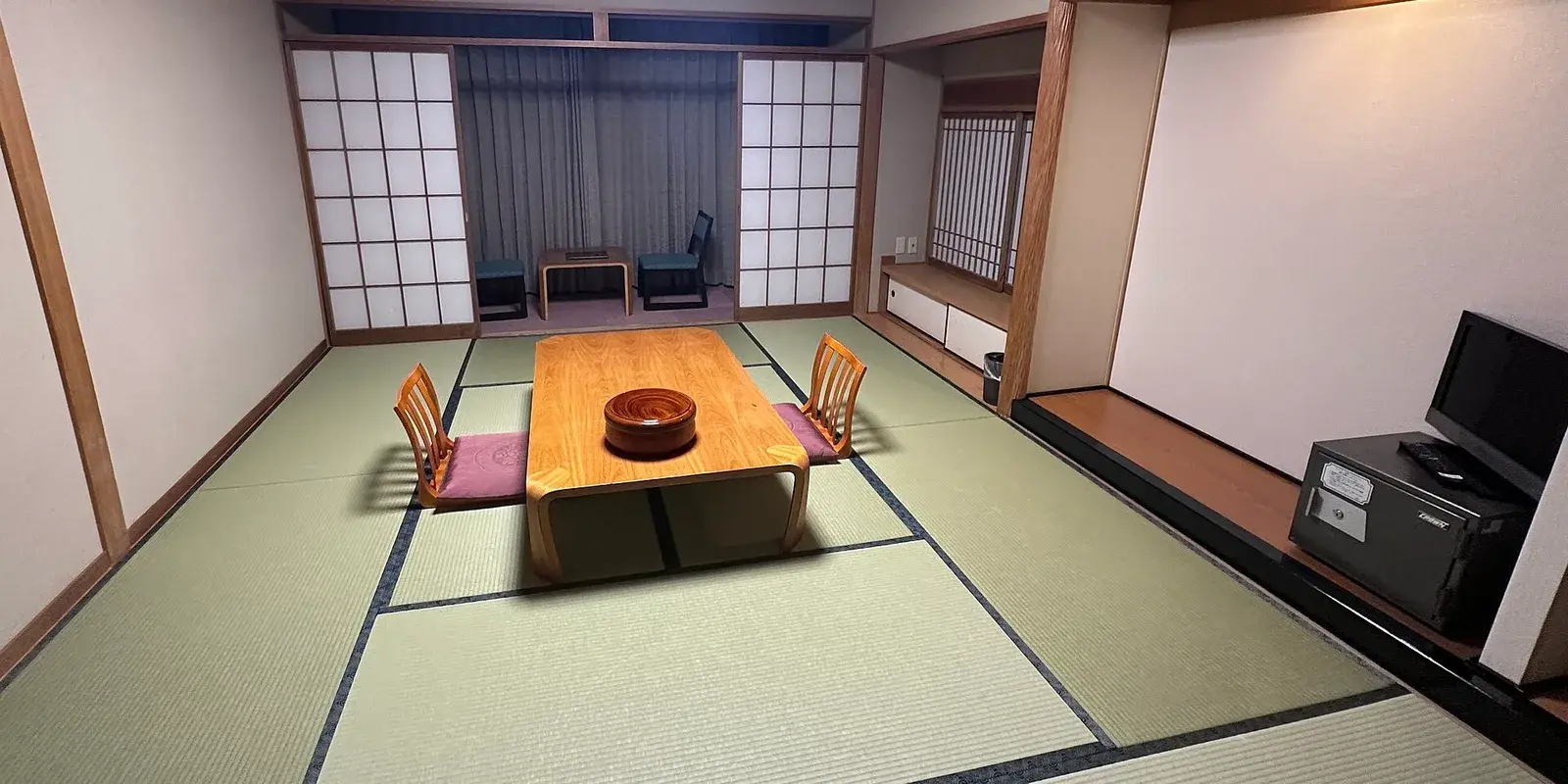A bright, traditional ryokan guest room with green tatami mats, a low wooden table flanked by legless zaisu chairs, paper shoji screens, and a small tea nook by the window