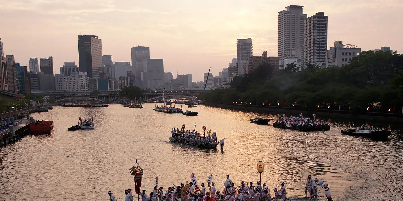 Lantern-lit boats of the Tenjin Matsuri funatogyo procession cross the Okawa River at dusk with the Osaka skyline behind