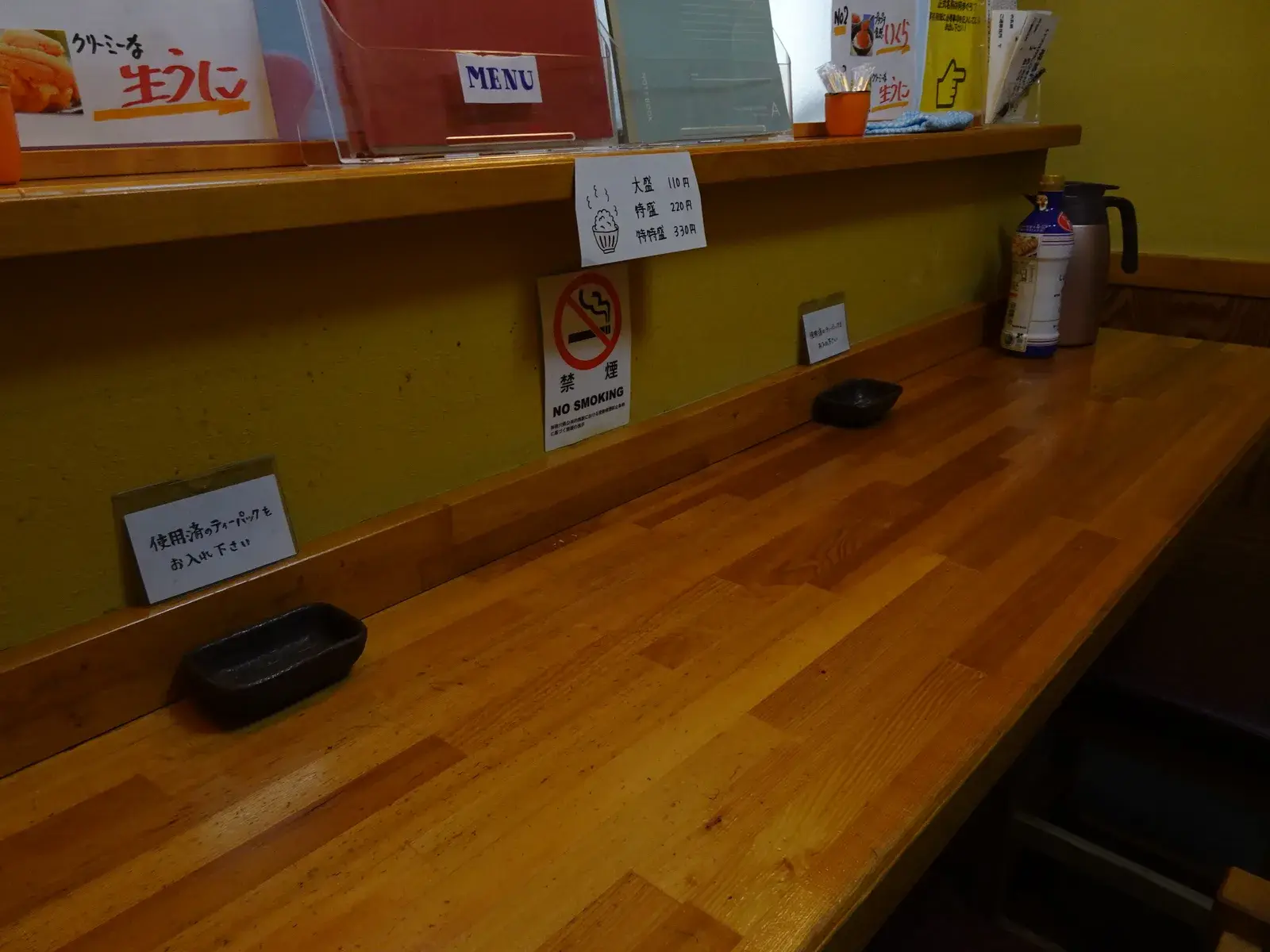 The empty wooden counter of a small seafood shop in Kawasaki, its seats unoccupied and the hot water flask waiting at the far end