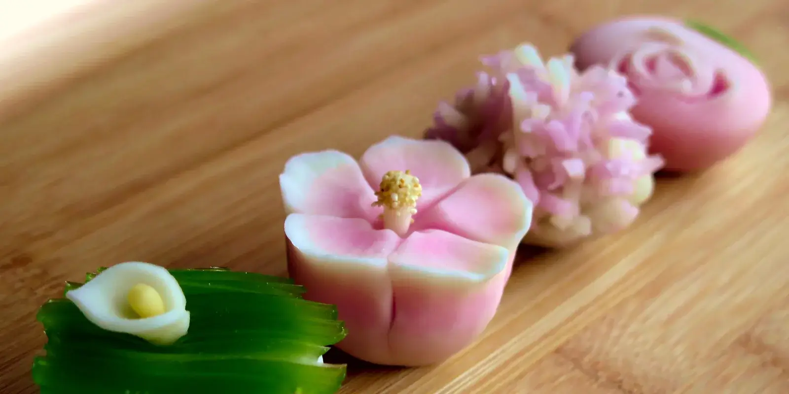 Three artisanal Japanese seasonal wagashi sweets shaped like flowers arranged on a warm wooden board