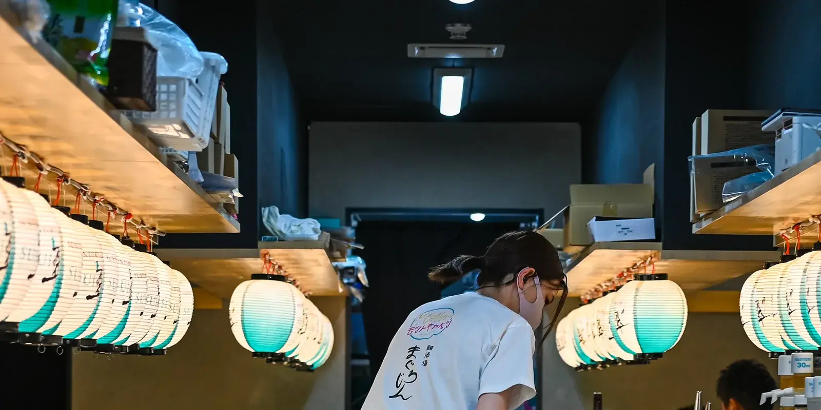 An izakaya staff member, seen from behind in a shop T-shirt, works in a narrow corridor lined on both sides with glowing green-and-white paper lanterns — the kind of warm Japanese service setting where a quick "sumimasen" calls or thanks the staff.