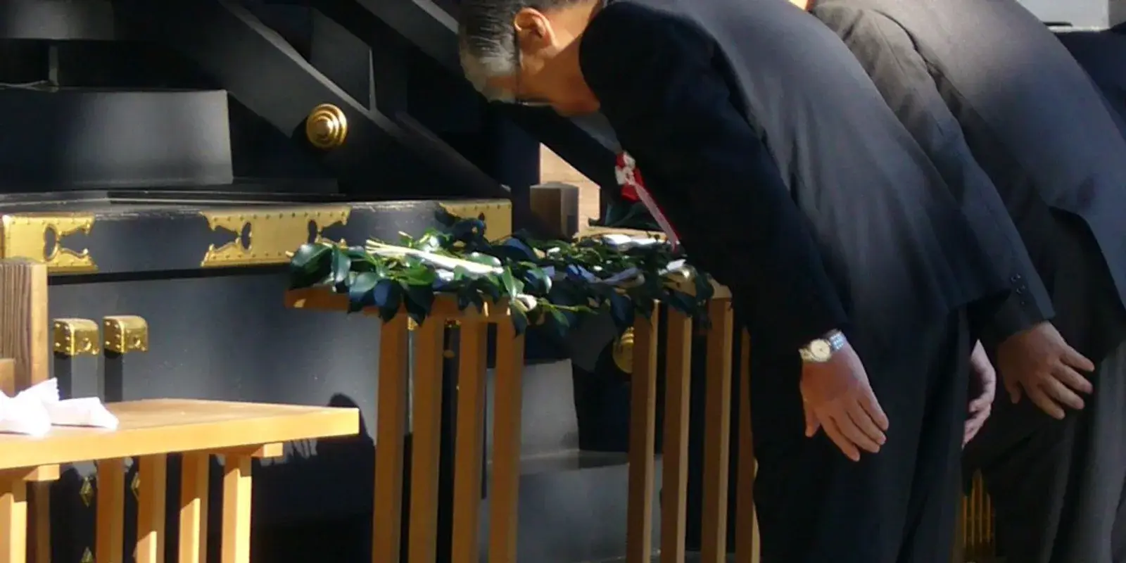Adult men in dark suits bow deeply in a row toward a shrine offering box draped with a sacred sakaki garland, hands on their knees and heads lowered