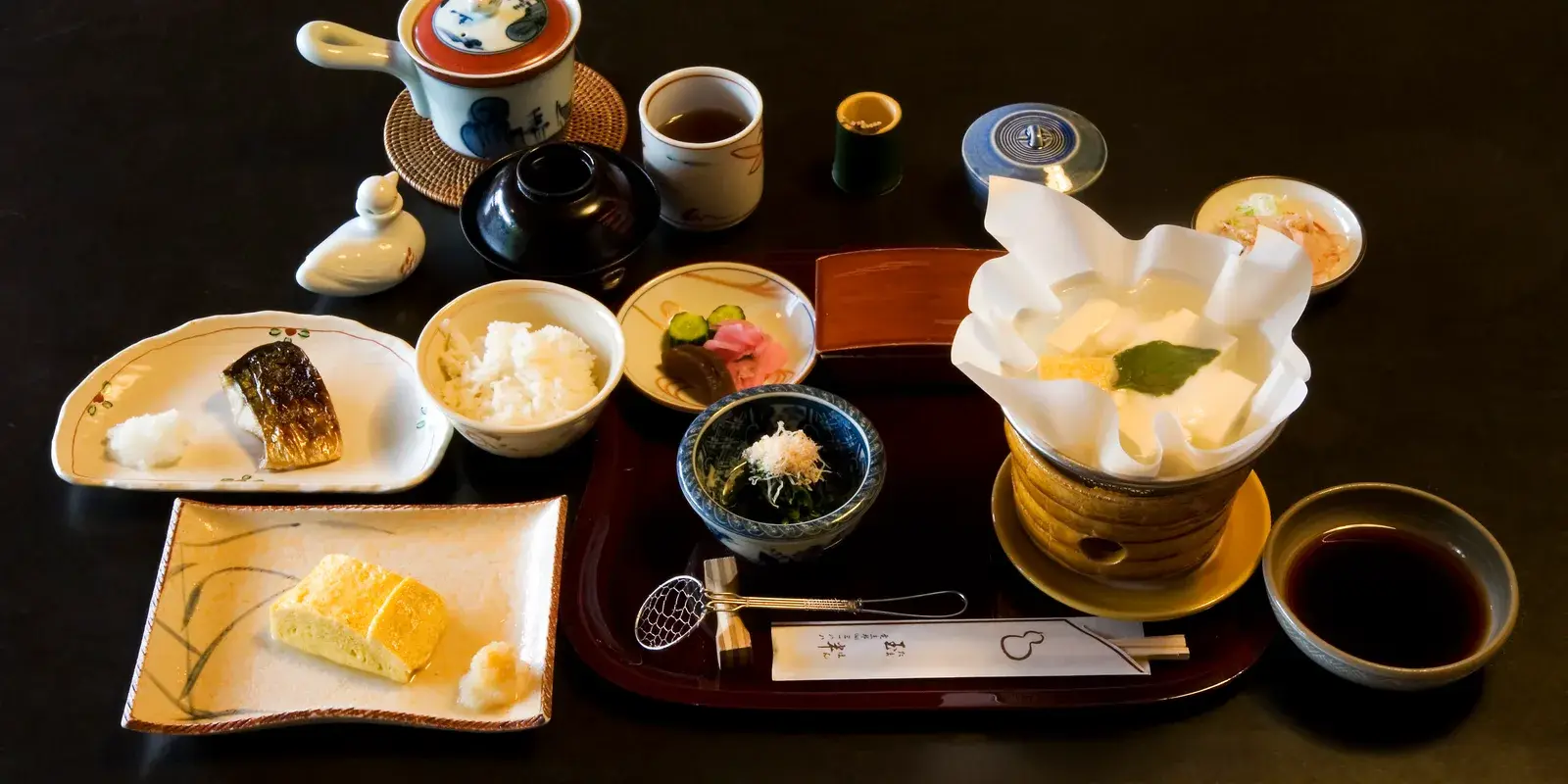 A traditional Japanese breakfast set on a dark table: grilled mackerel, a bowl of rice, dashimaki egg, yudofu simmering in a paper pot, pickles, soy sauce, and green tea, arranged on a lacquer tray