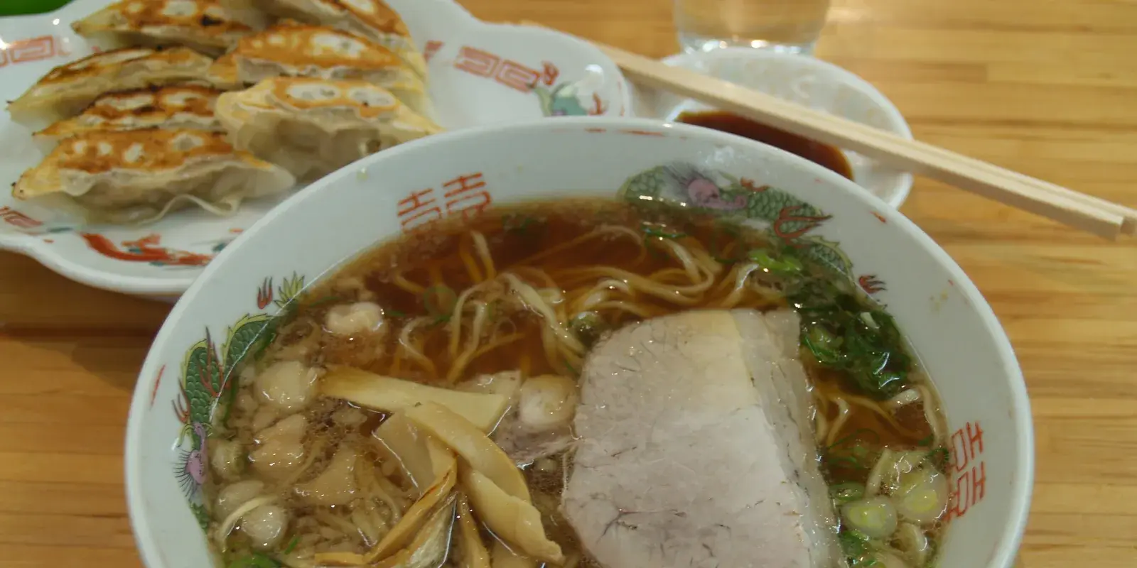 A bowl of Onomichi-style shoyu ramen with chashu pork, menma, and scallions, served with a plate of gyoza and chopsticks on a wooden table